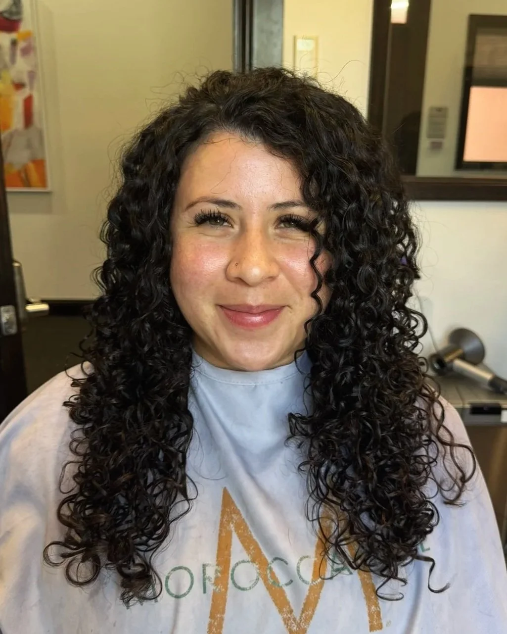 A woman with long, curly dark hair and a light complexion smiling at the camera. She is seated in a hair salon wearing a cape with a logo, and there are salon mirrors and equipment in the background.