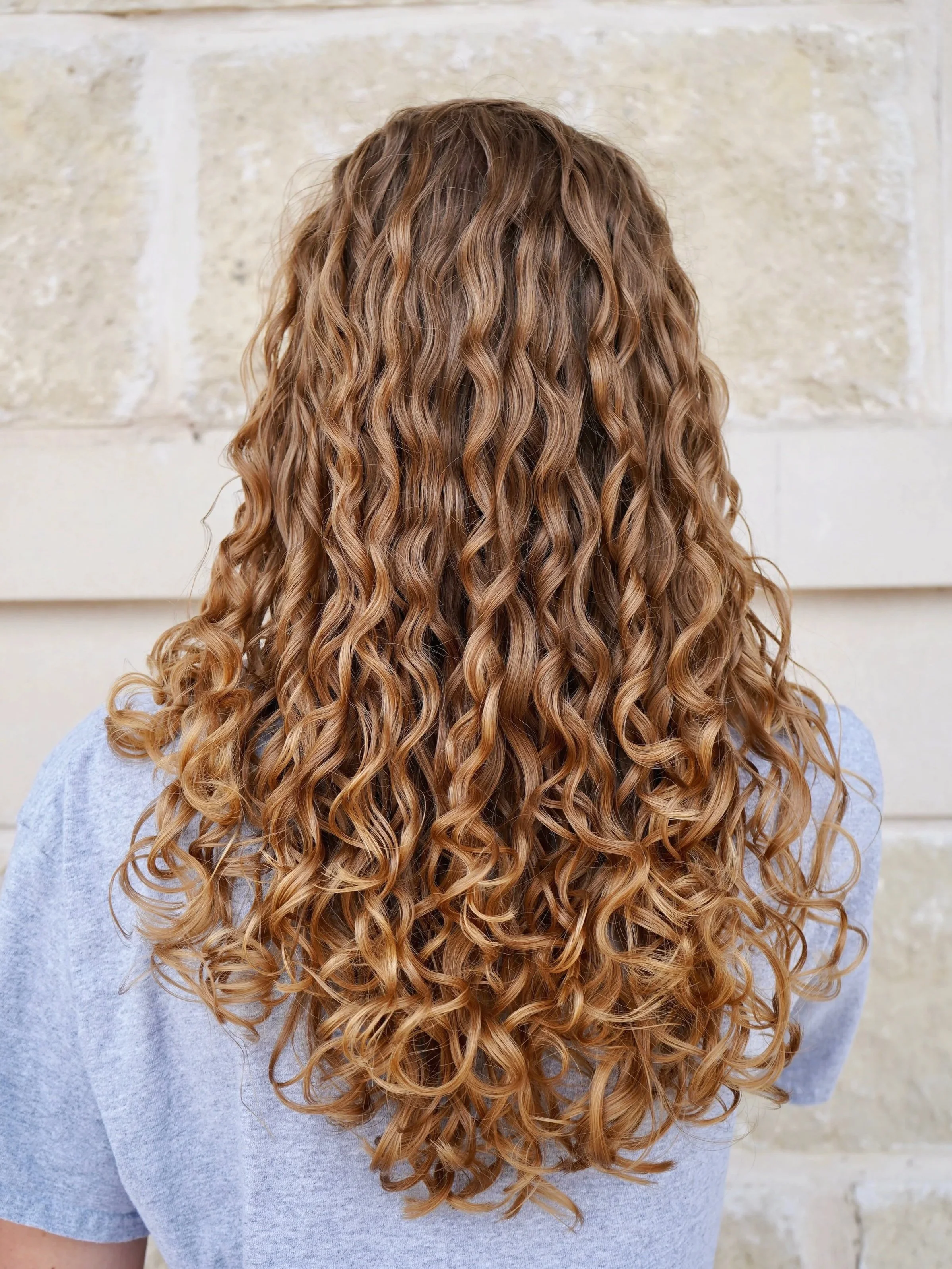 Back view of a person with long, curly, light brown hair wearing a light purple shirt, standing in front of a stone wall.
