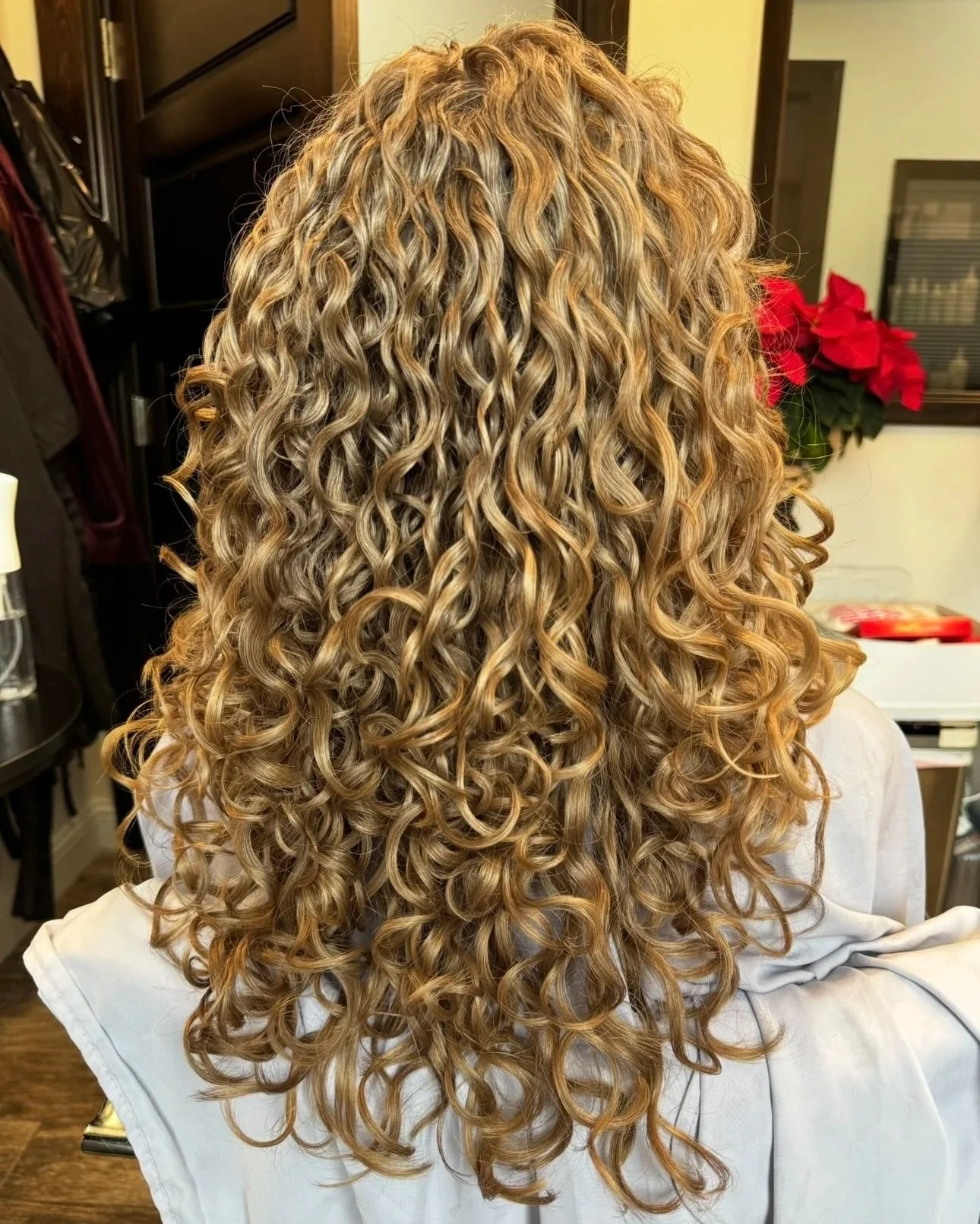 Back of a woman with long, curly blonde hair in tight ringlet curls, seated in a salon chair.