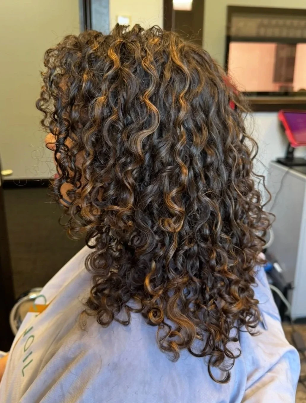 Person with long, curly hair sitting in a salon chair, facing away, with a salon station and mirror in the background.