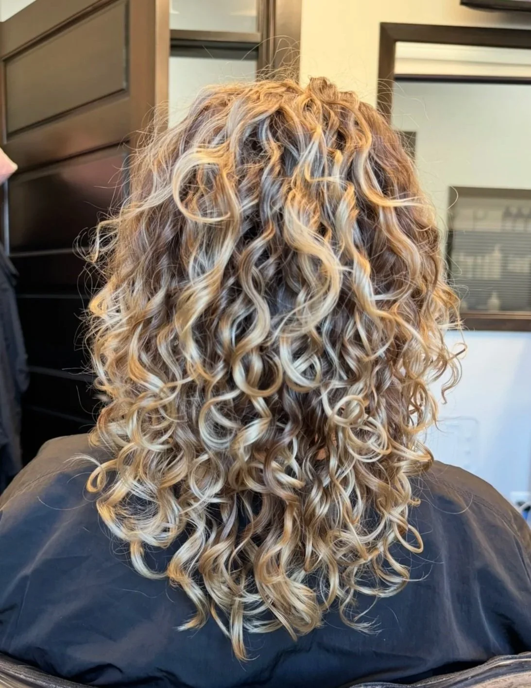 Back view of a person with long, curly blonde hair sitting in a salon chair.