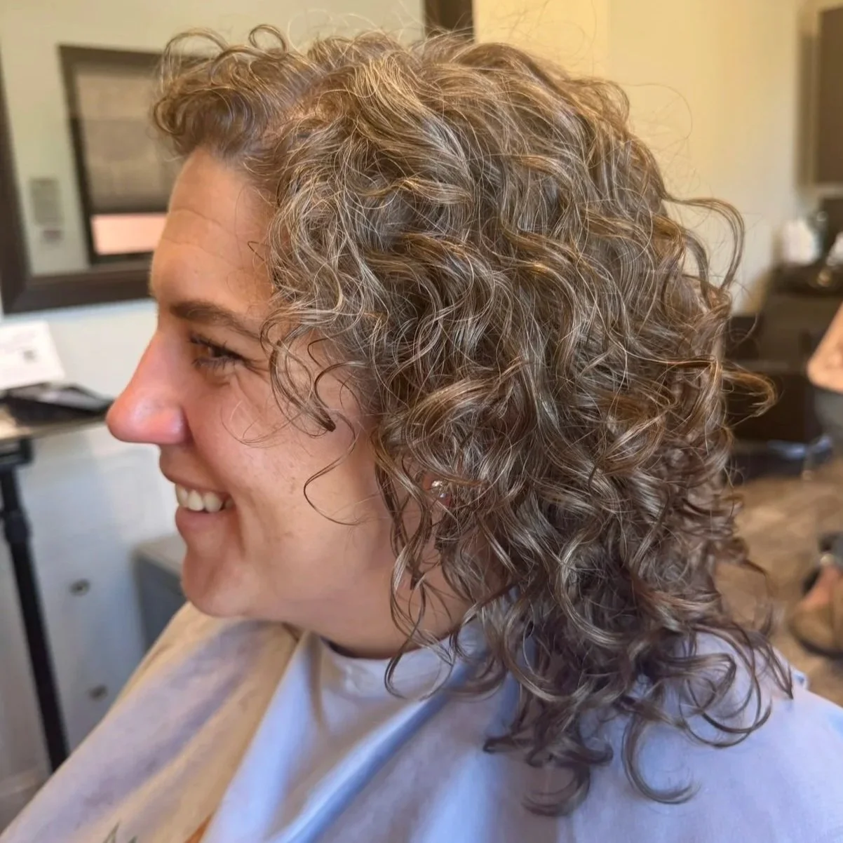 A woman with curly, brown hair in a salon chair, smiling, wearing a cape. In the background, a person stands near a desk and a television.