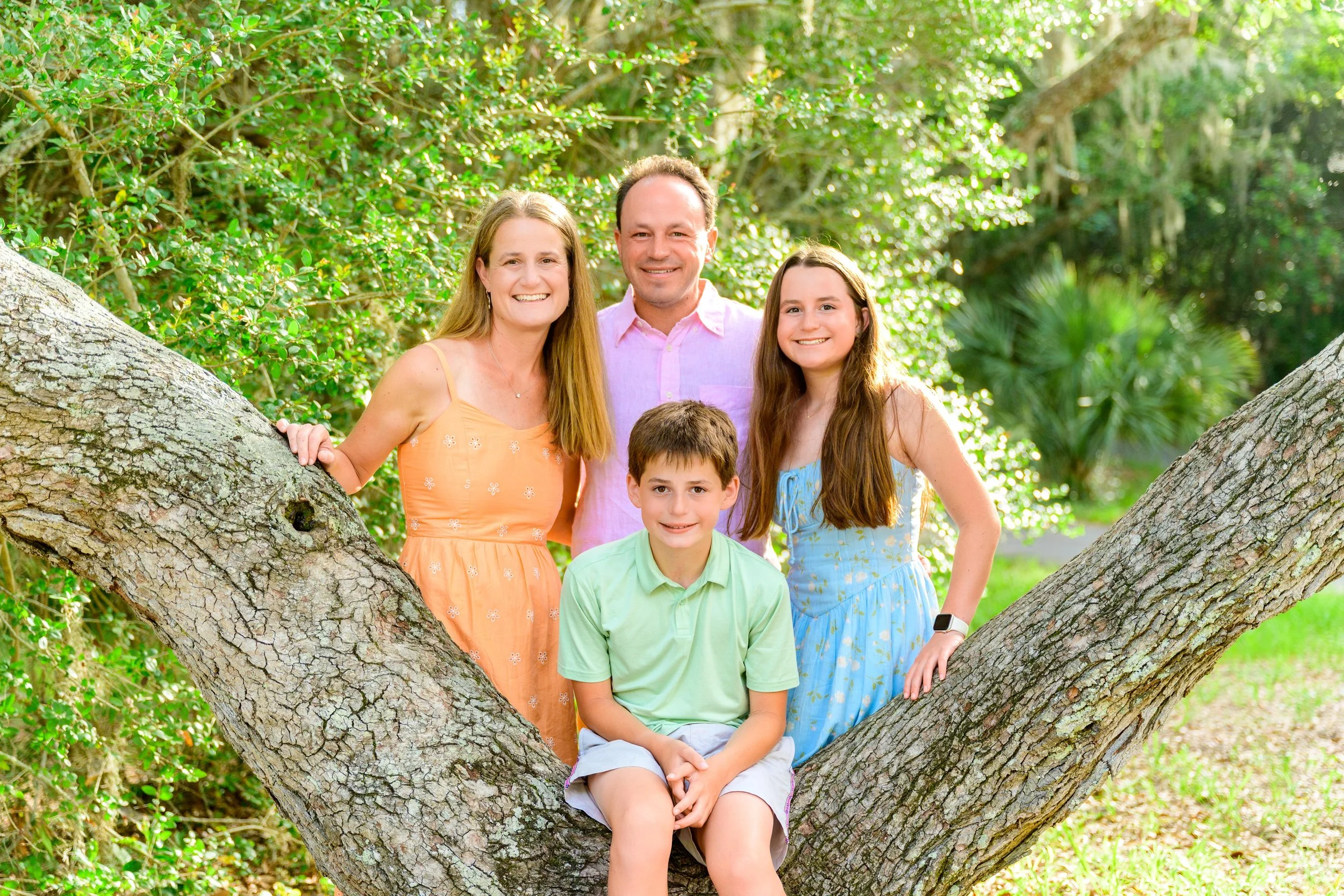 Matt's family posing outdoors on a large tree with lush green foliage in the background, smiling at the camera.