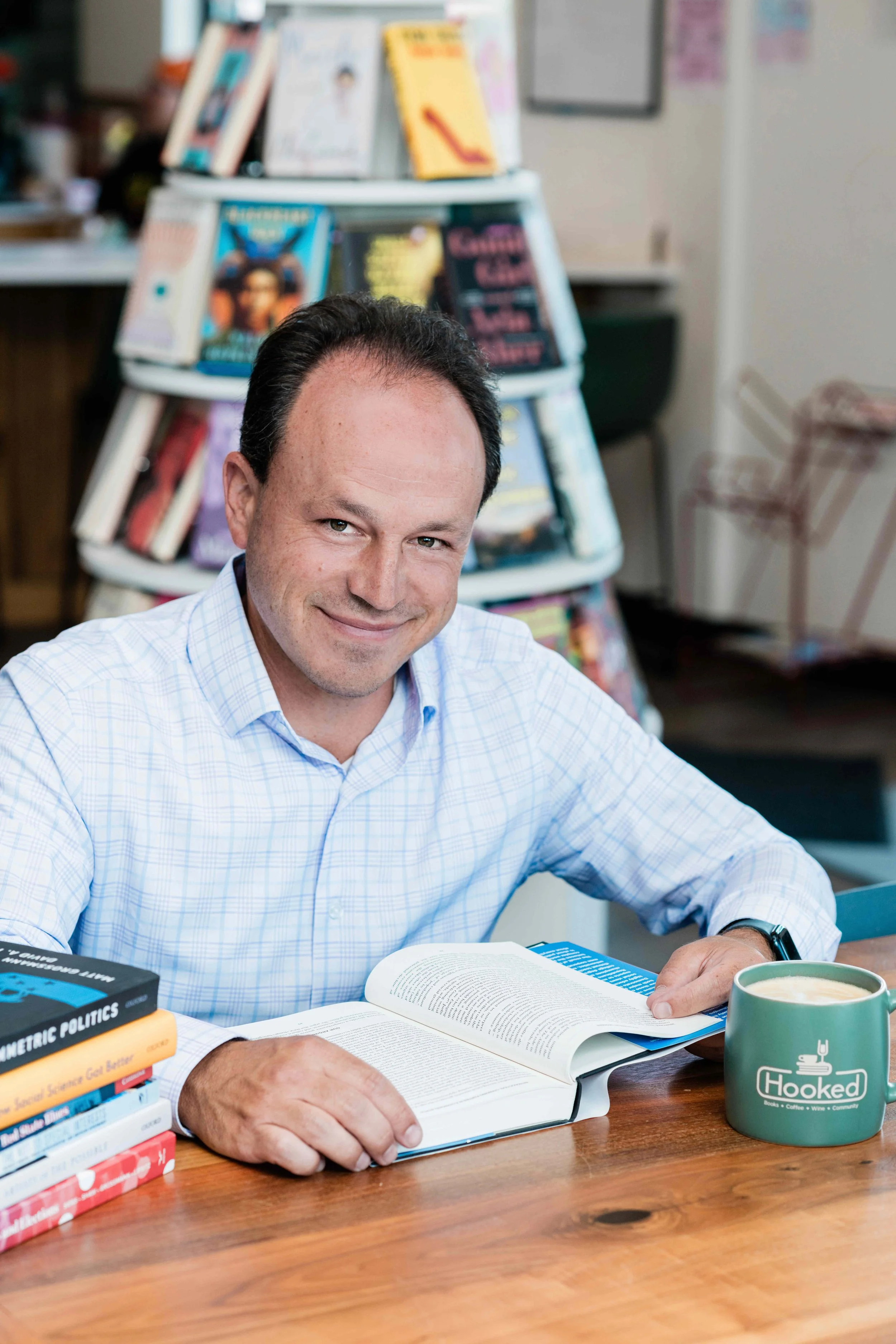 Matt sitting at a wooden table with an open book, a stack of books, a green mug, smiling at the camera in a bookstore.