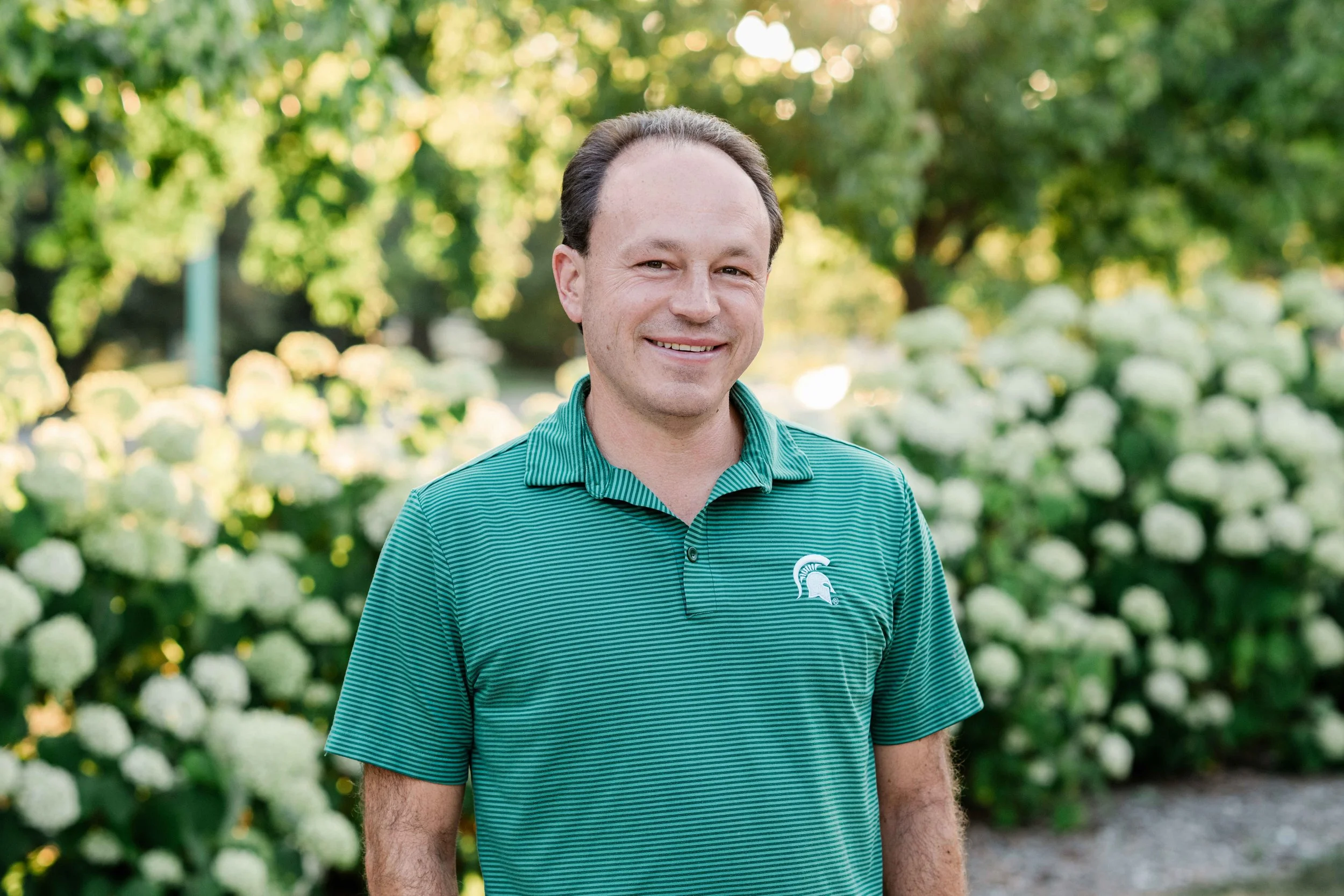 Matt Grossmann in a green and black striped polo shirt with a Michigan State Spartans logo, standing outdoors in front of white hydrangea flowers and green trees.