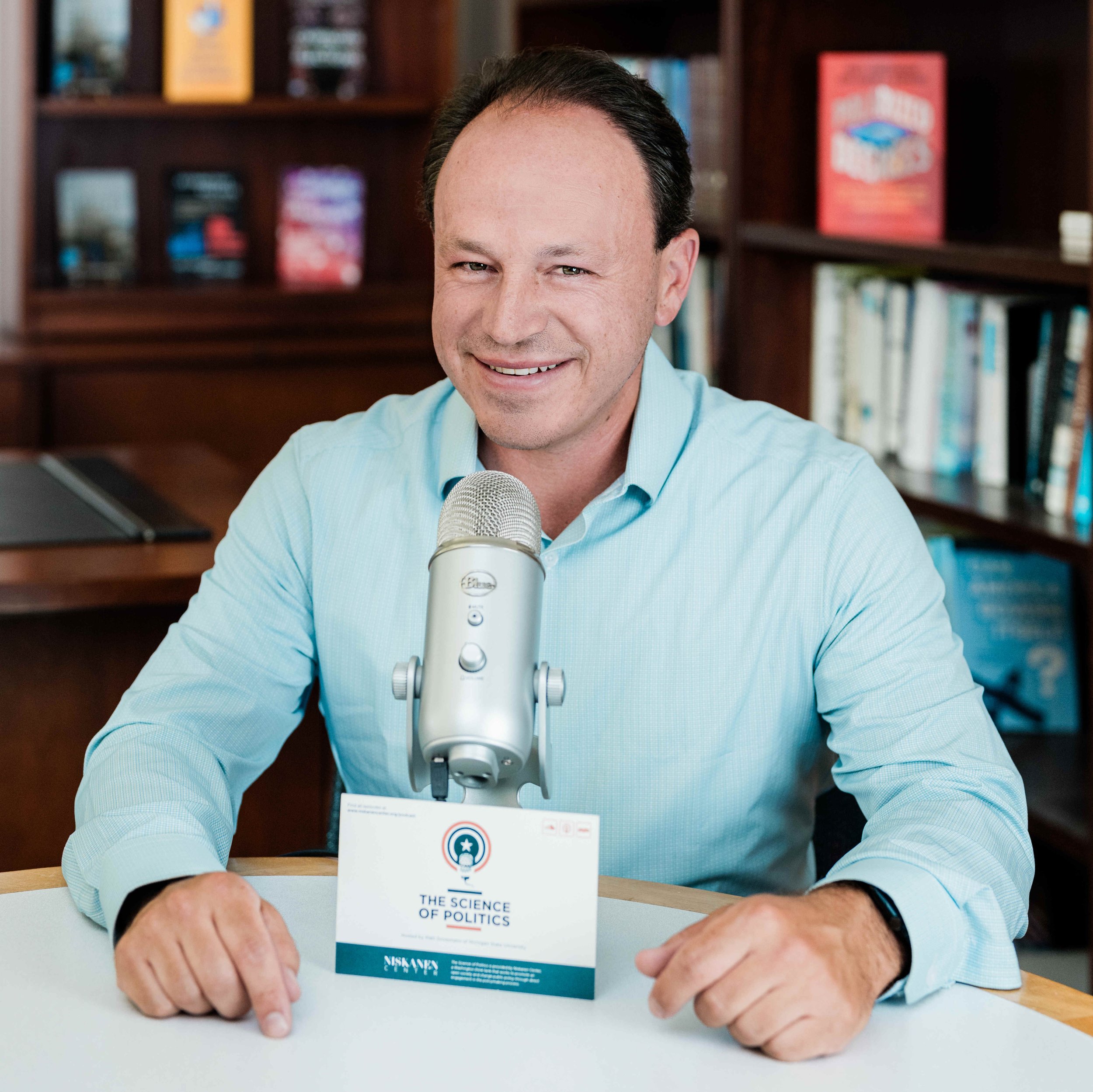 Matt Grossmann sitting at a desk, smiling, with a microphone in front of him, in a room with bookshelves in the background.