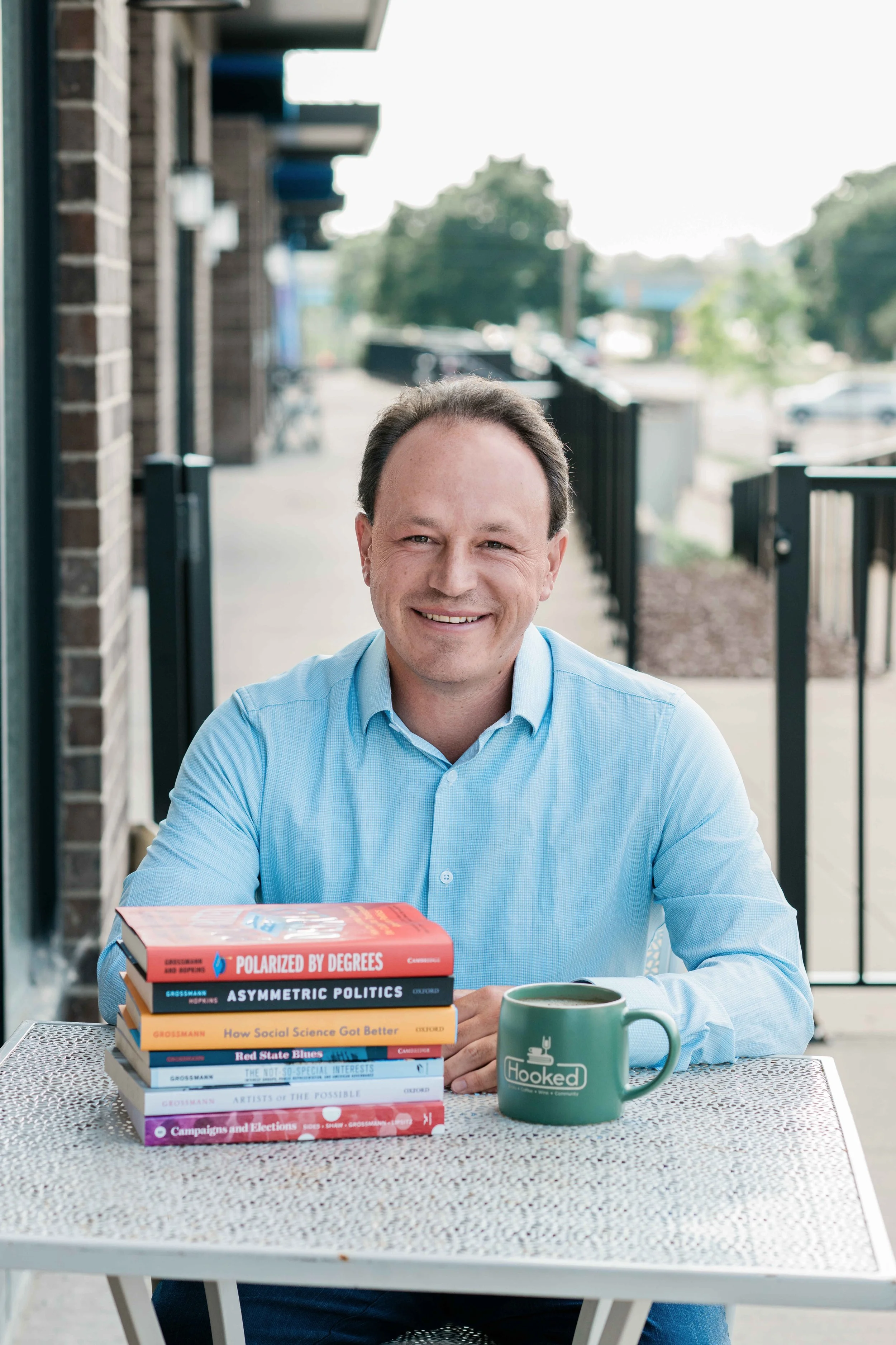 Matt smiling in a light blue shirt sitting at a table outdoors, with a stack of books and a green mug labeled 'Hooked' in front of him.