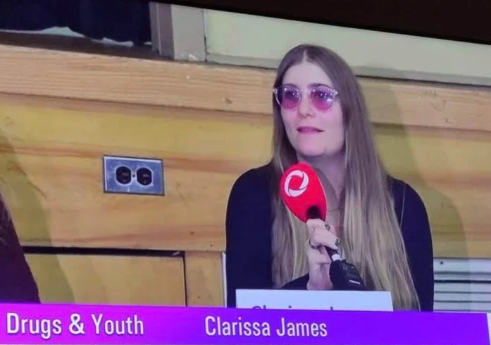 A woman with long blonde hair and pink sunglasses sitting in front of a wooden panel. She is holding a red microphone with a white logo. There is a purple banner at the bottom that says "Drugs & Youth" and "Clarissa James."