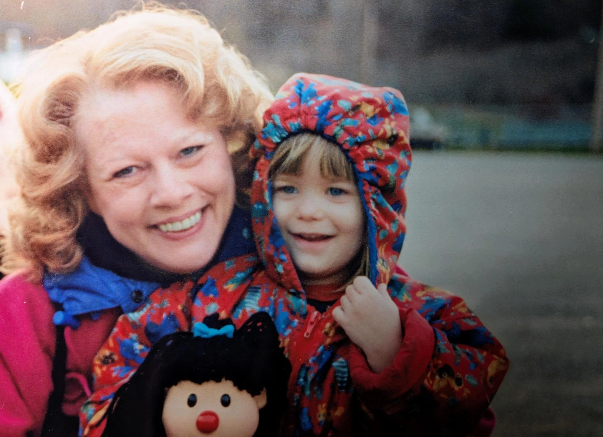 A woman with curly blonde hair smiling and a young girl with a colorful hooded jacket, also smiling, holding a doll with black hair and a red nose.