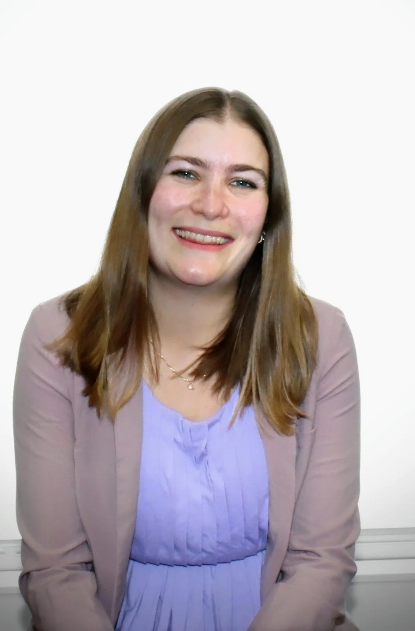 A young woman with straight, shoulder-length brown hair, smiling, wearing a lavender blouse and a beige blazer, sitting in front of a plain white background.
