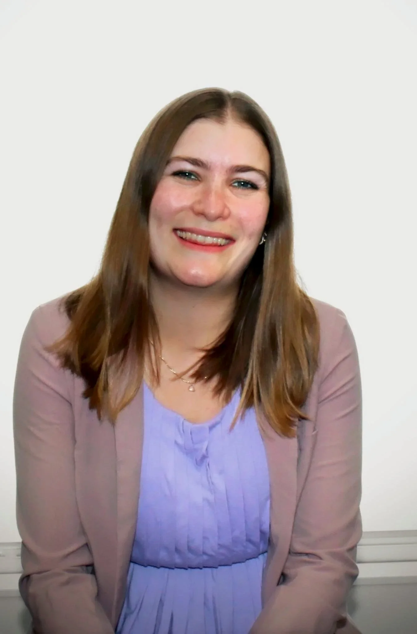 A woman with shoulder-length brown hair, wearing a lavender blouse and a beige blazer, smiling against a plain white background.