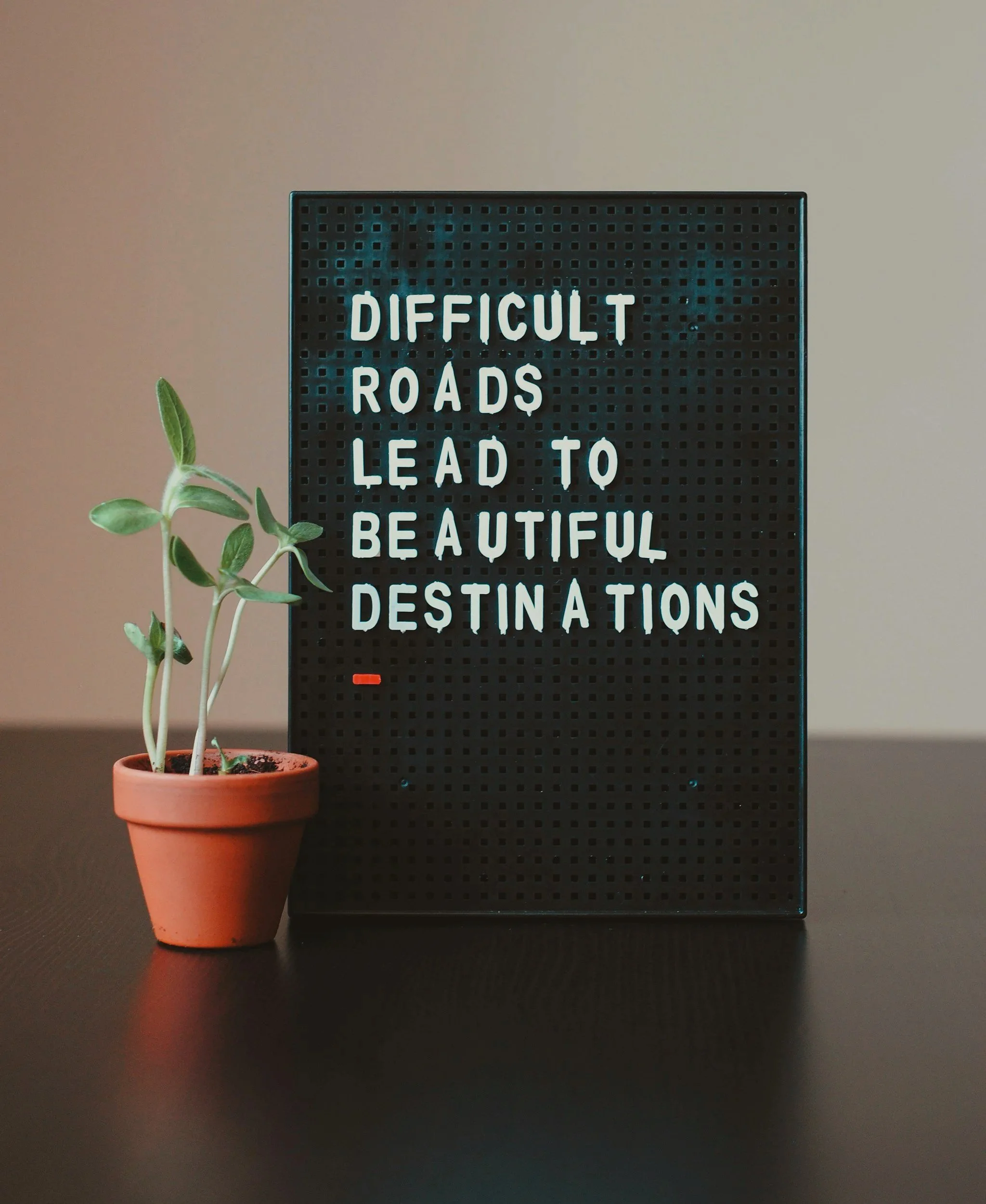 A black light-up letter board displaying the quote "Difficult Roads Lead to Beautiful Destinations" next to a small potted green plant on a black surface.