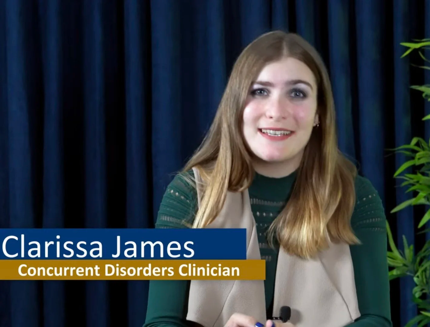 A woman with long reddish hair sitting in front of a dark blue curtain, wearing a green top and beige vest, smiling at the camera. Text overlay: "Clarissa James, Concurrent Disorders Clinician."