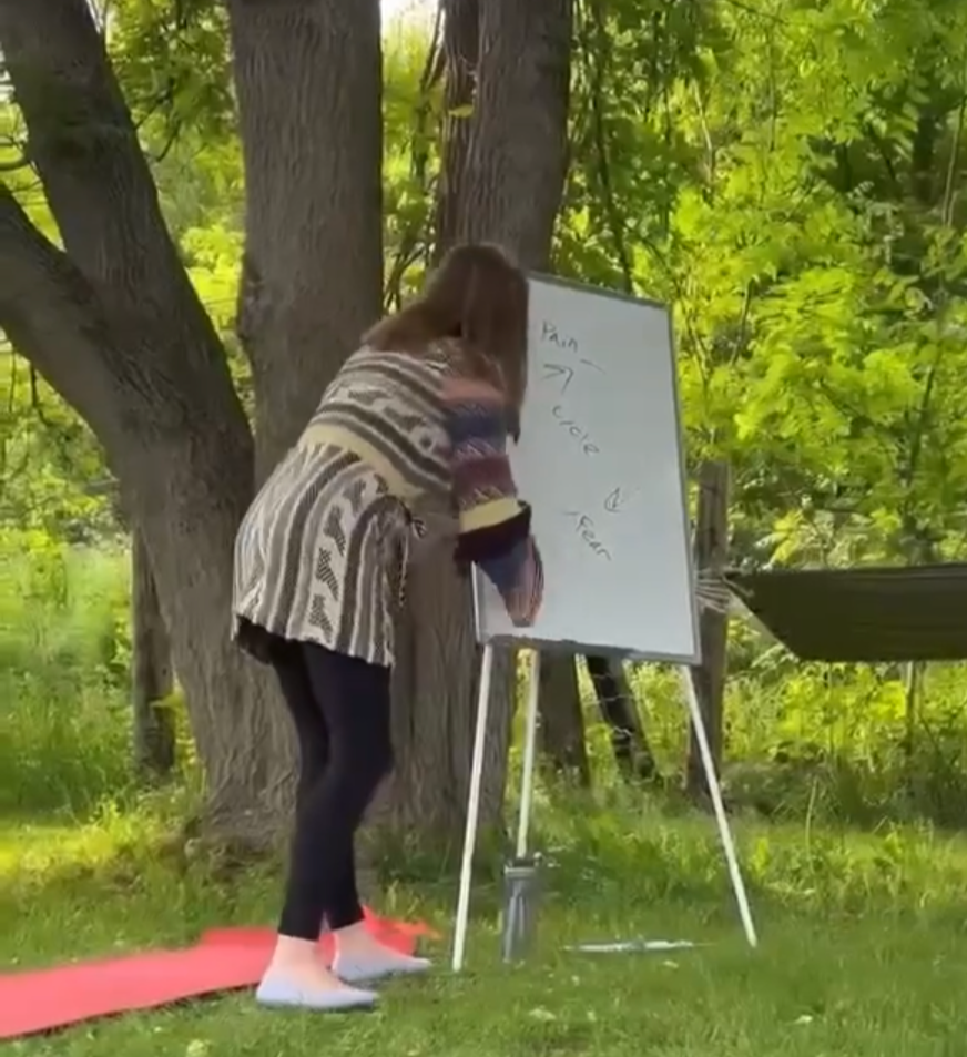 A woman standing outdoors on a red yoga mat, writing on a whiteboard with a tree and green foliage in the background.