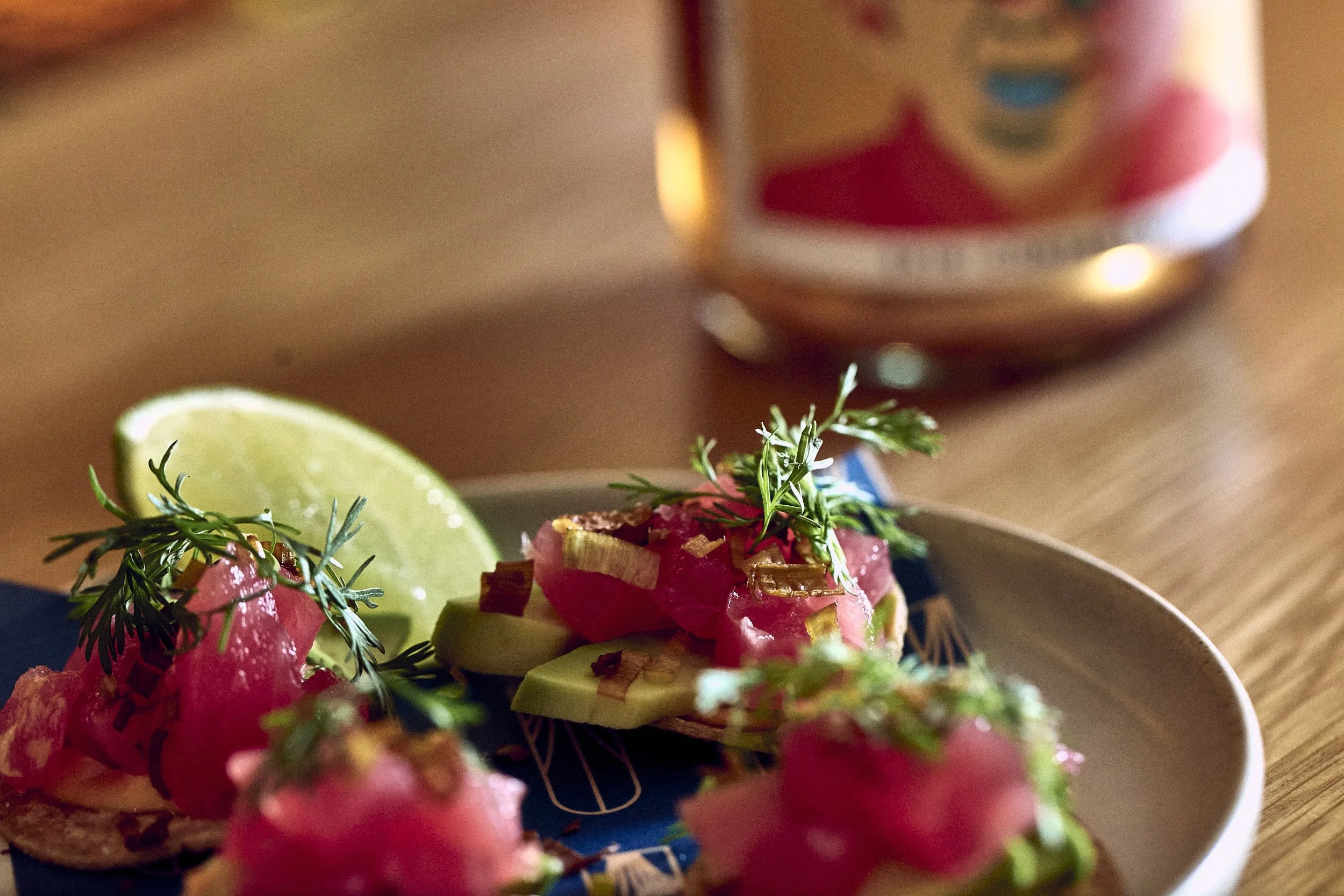 Close-up of a dish with diced pink meat, garnished with herbs, lime wedge, and crispy toppings, with a bottle of ketchup in the background.