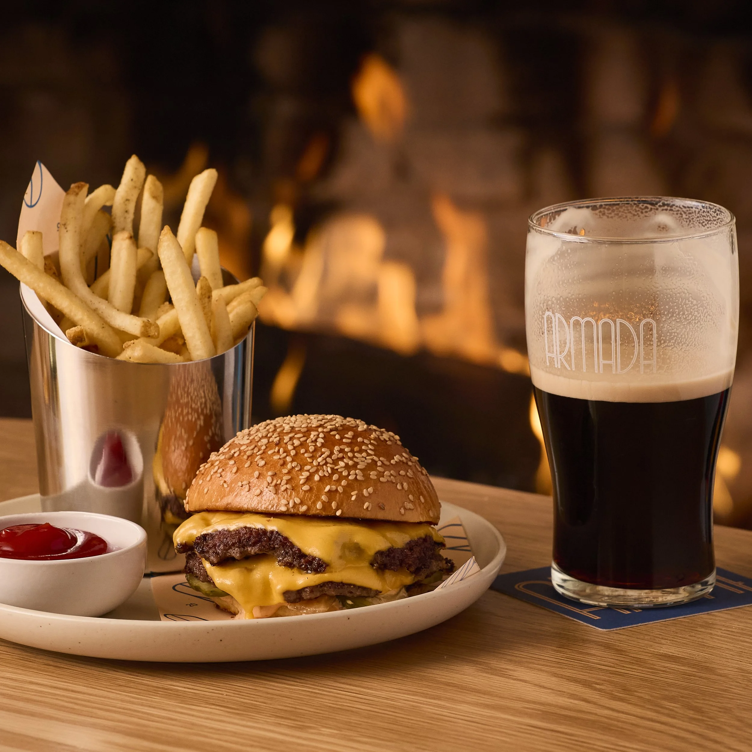 A cheeseburger with a sesame seed bun, melted cheese, and beef patties on a white plate, alongside a small bowl of ketchup. Two glasses of dark soda with foam are in the background, with a blurry fire or warm light source behind them.