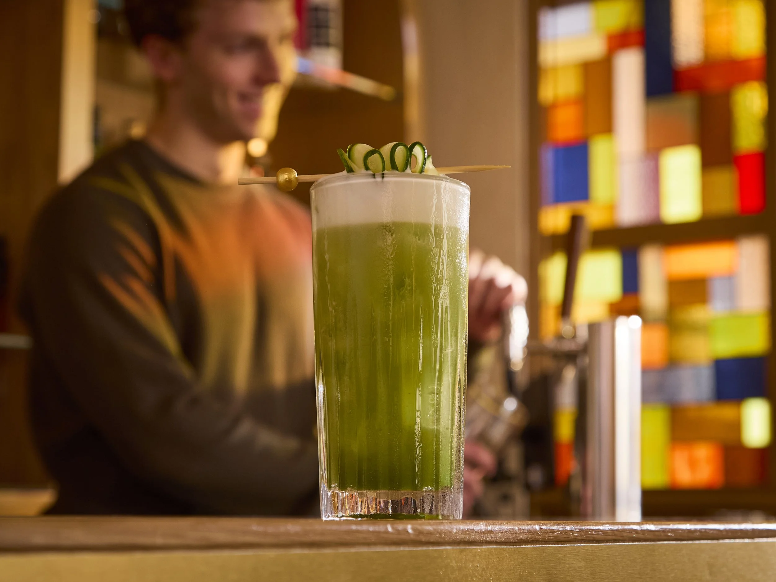 A tall glass filled with a green beverage, garnished with cucumber spirals and skewered with a decorative stick, on a wooden surface with a blurred background of a person and colorful shelves.