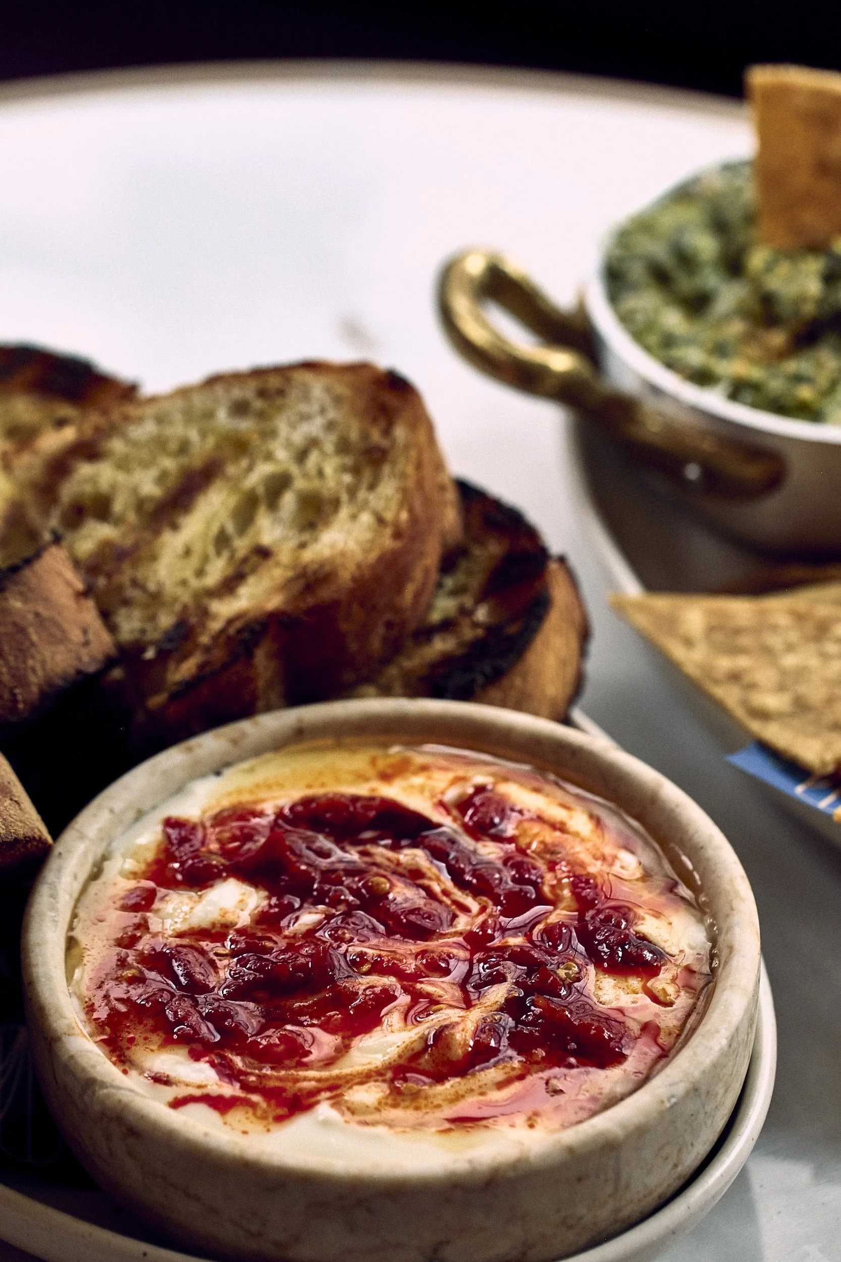 A bowl of cranberry sauce. In the background, there are slices of bread and a cup of spinach and cheese dip or spread.