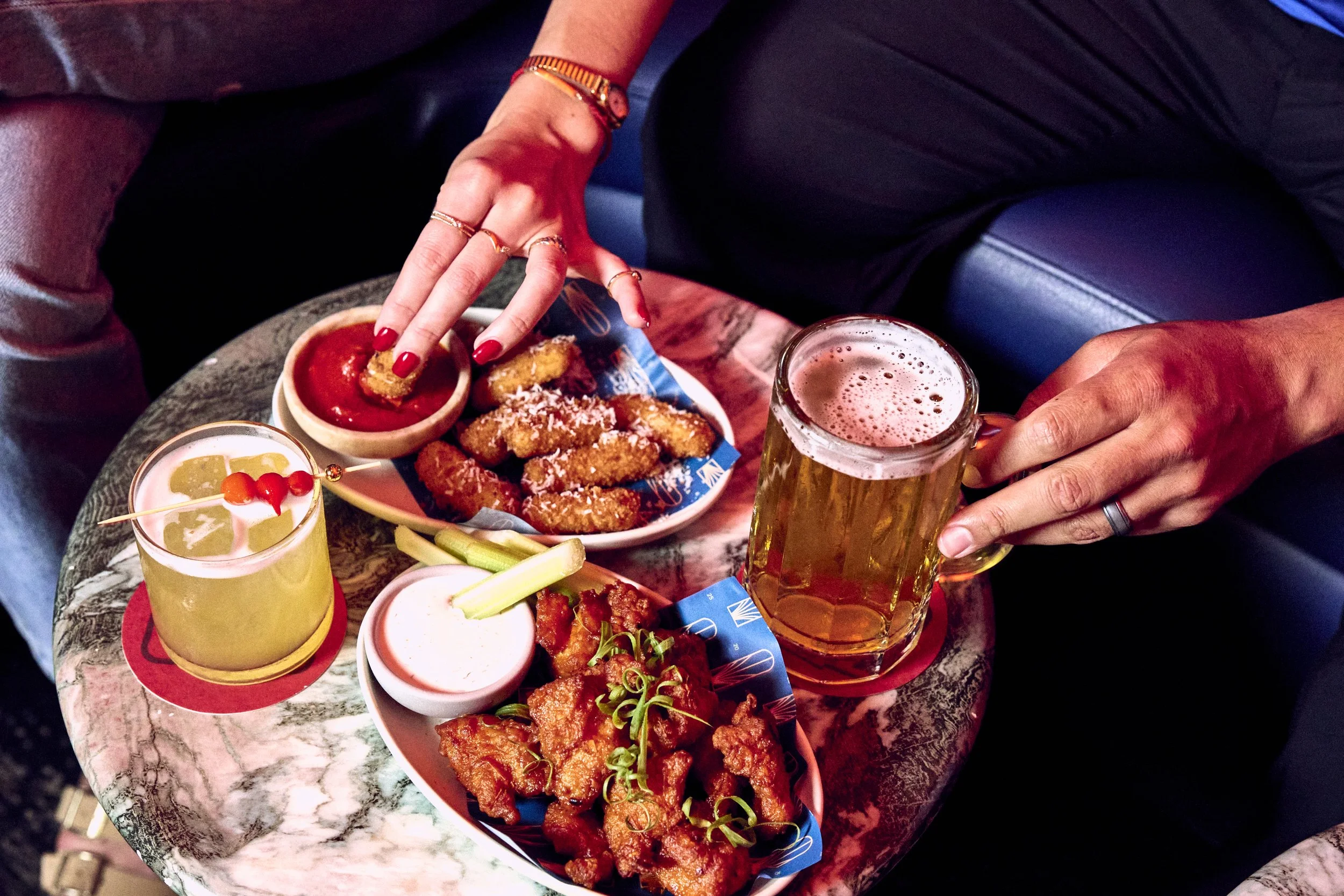 Person reaching for fried appetizers on a table with beers and drinks, including a bowl of fried chicken, dipping sauces, celery sticks, and a cocktail with garnishes, on a marble table.