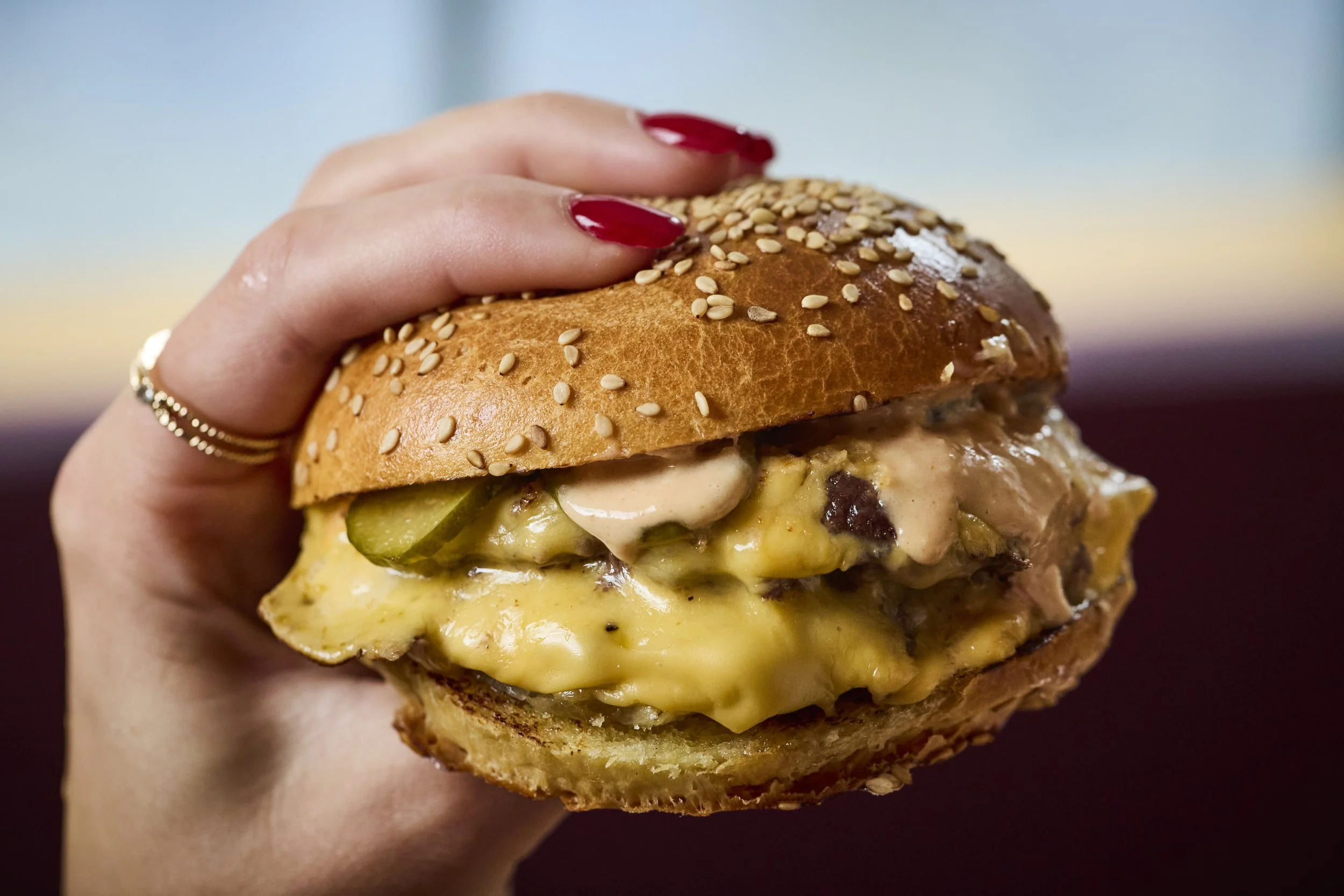 Close-up of a hand with red nail polish holding a cheeseburger with sesame seed bun, melted cheese, pickles, sauce, and a beef patty.
