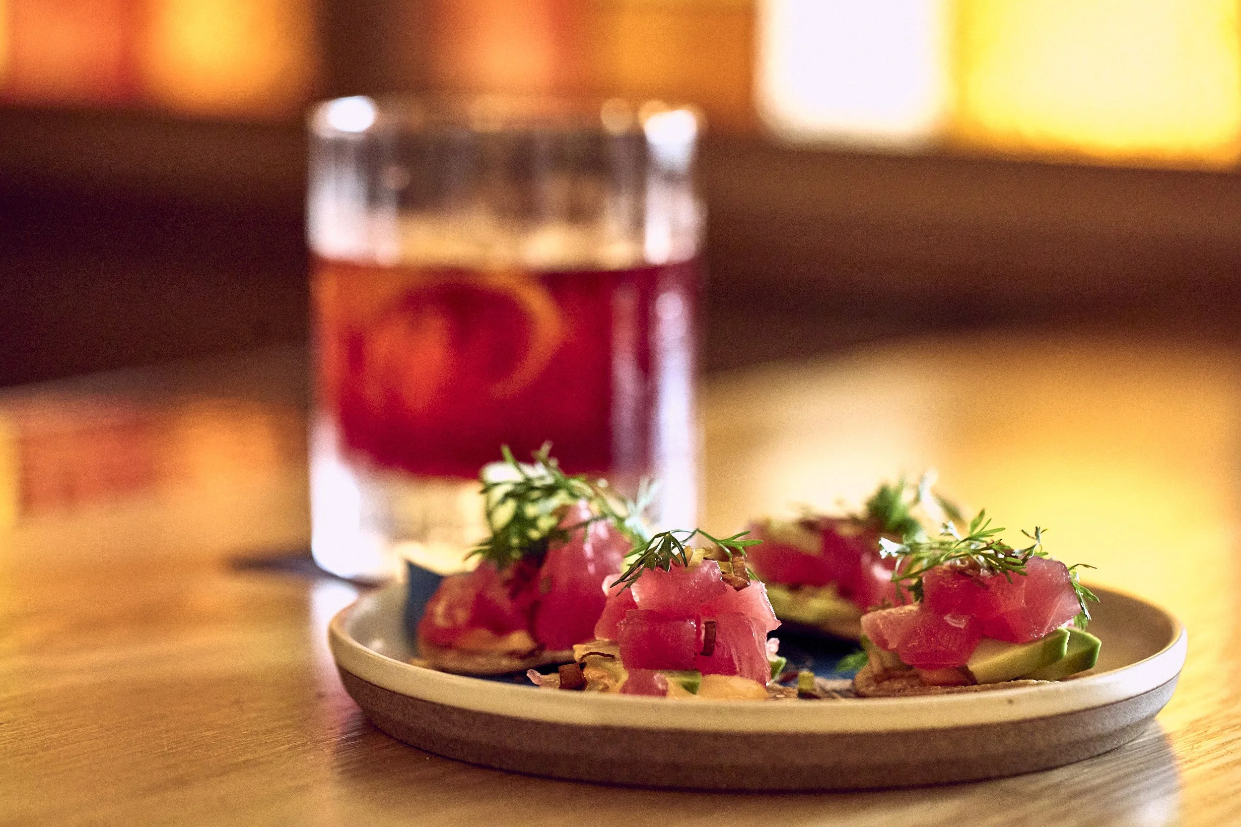 A small plate of food with sliced radishes and herbs on top, with a blurred drink in the background.