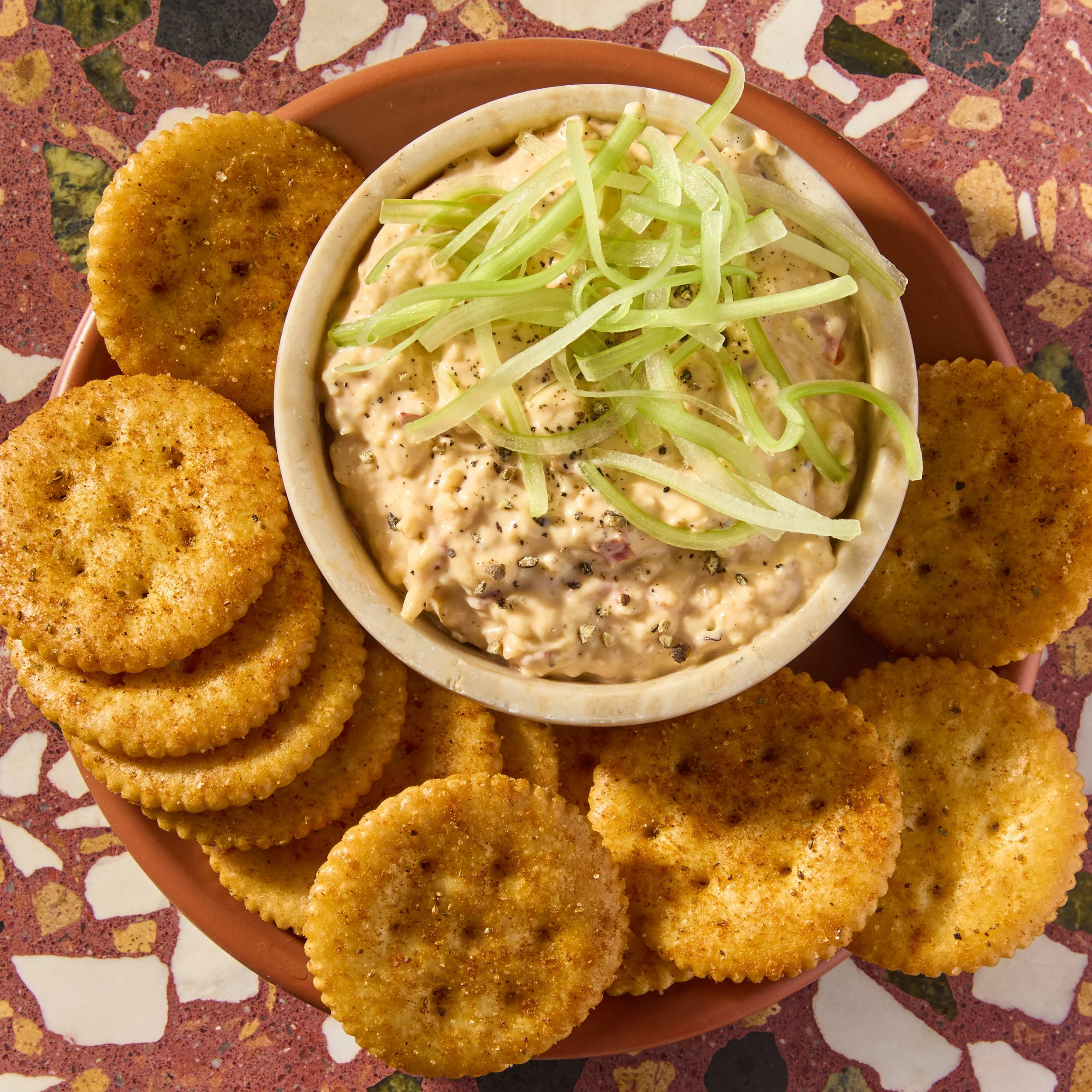 A round pink bowl containing breaded mozzarella sticks around a small bowl of creamy ranch dip topped with shredded lettuce and black pepper on a colorful speckled table.