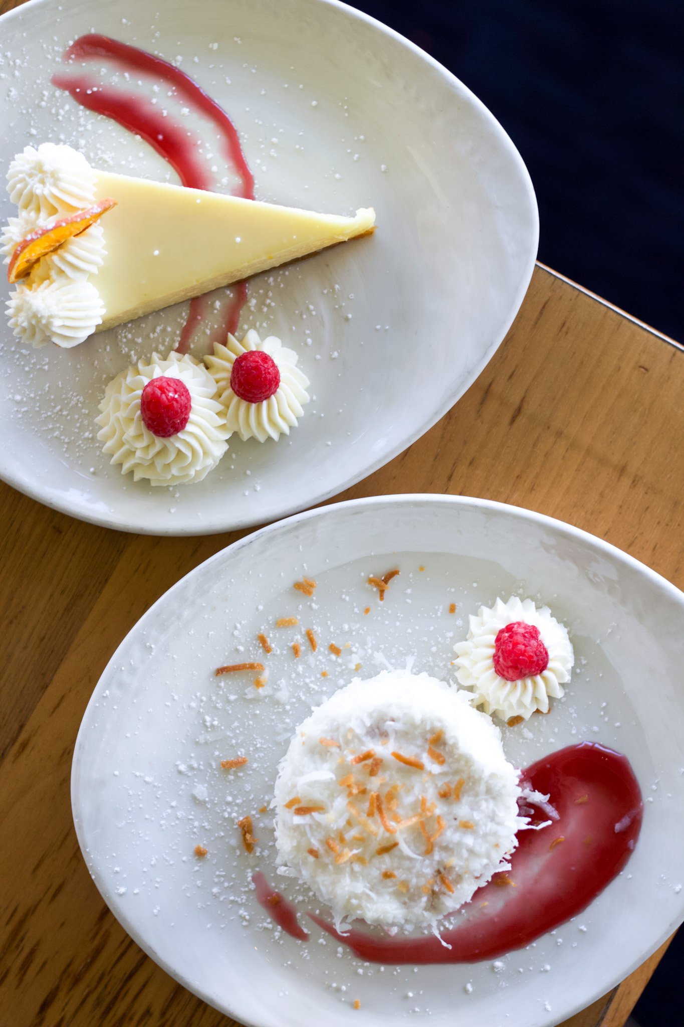 Two plates with desserts: a slice of cheesecake topped with whipped cream and raspberries, and a scoop of coconut ice cream with shredded coconut, whipped cream, and raspberry sauce.