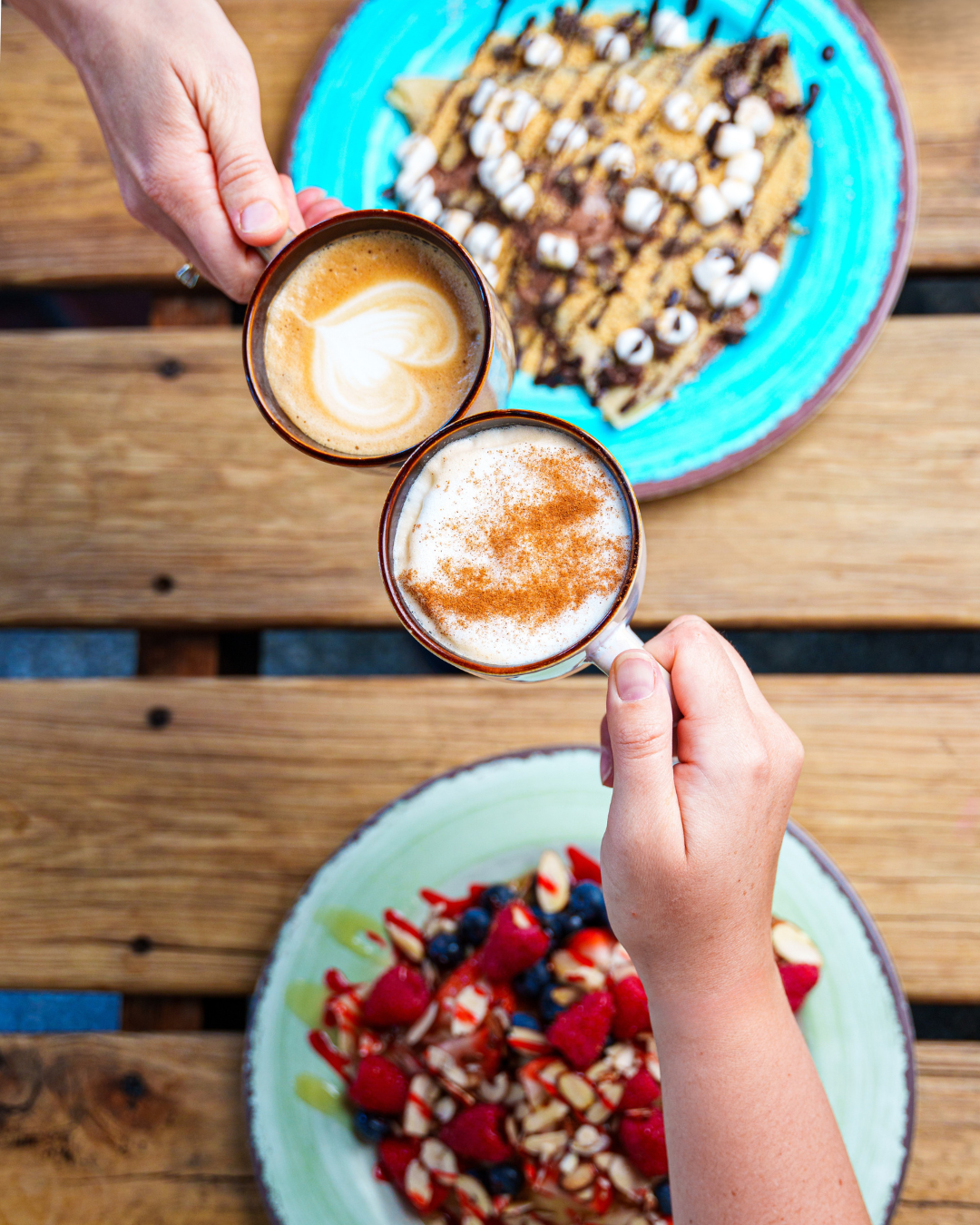 Two people holding coffee mugs with lattes over a wooden table with plates of waffles topped with marshmallows, chocolate, and whipped cream, and a berry dessert with strawberries, blueberries, and nuts.