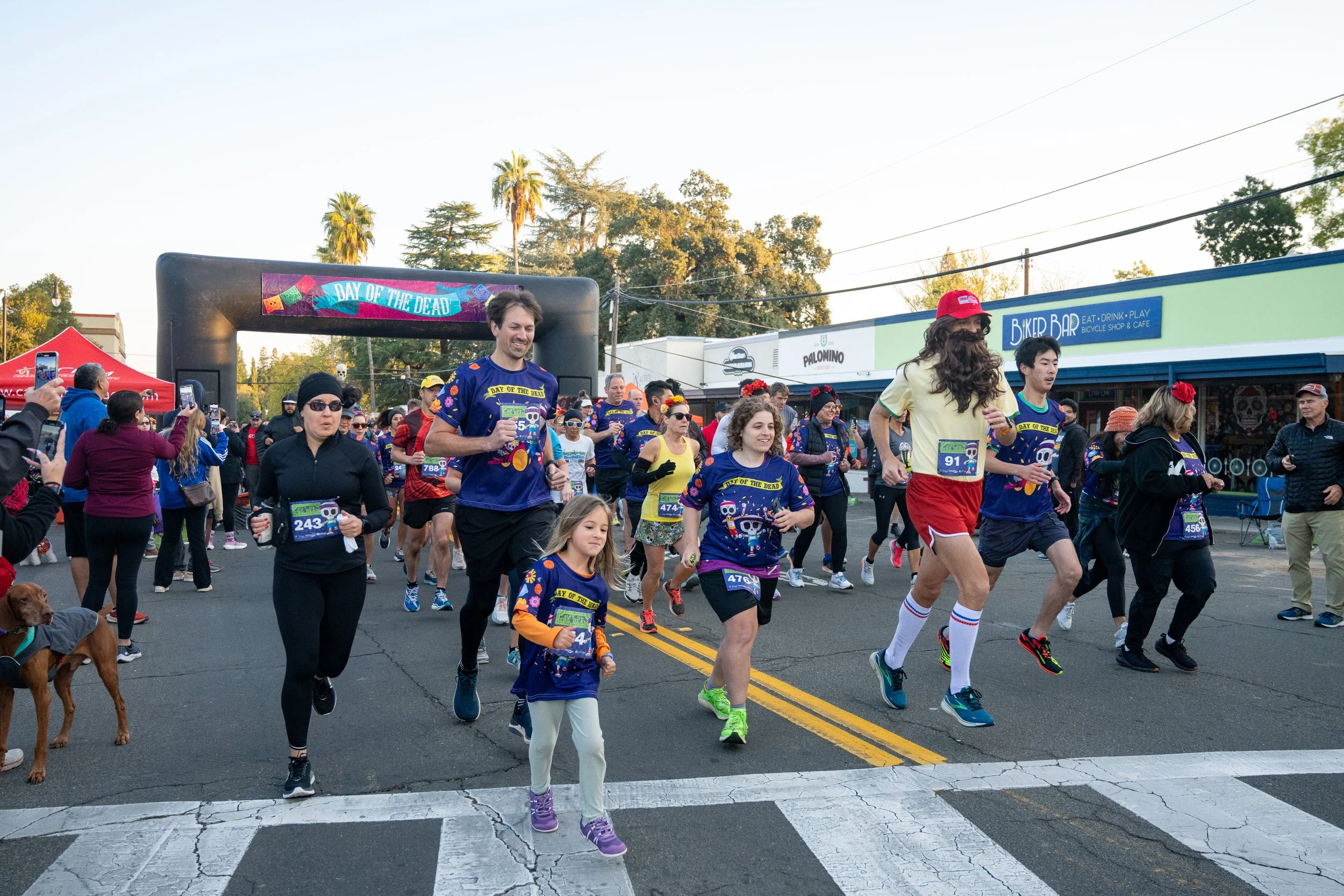 People participating in a race, many wearing themed shirts for a Día de los Muertos event, crossing a street with an arch that says 'Day of the Dead' and spectators taking photos.