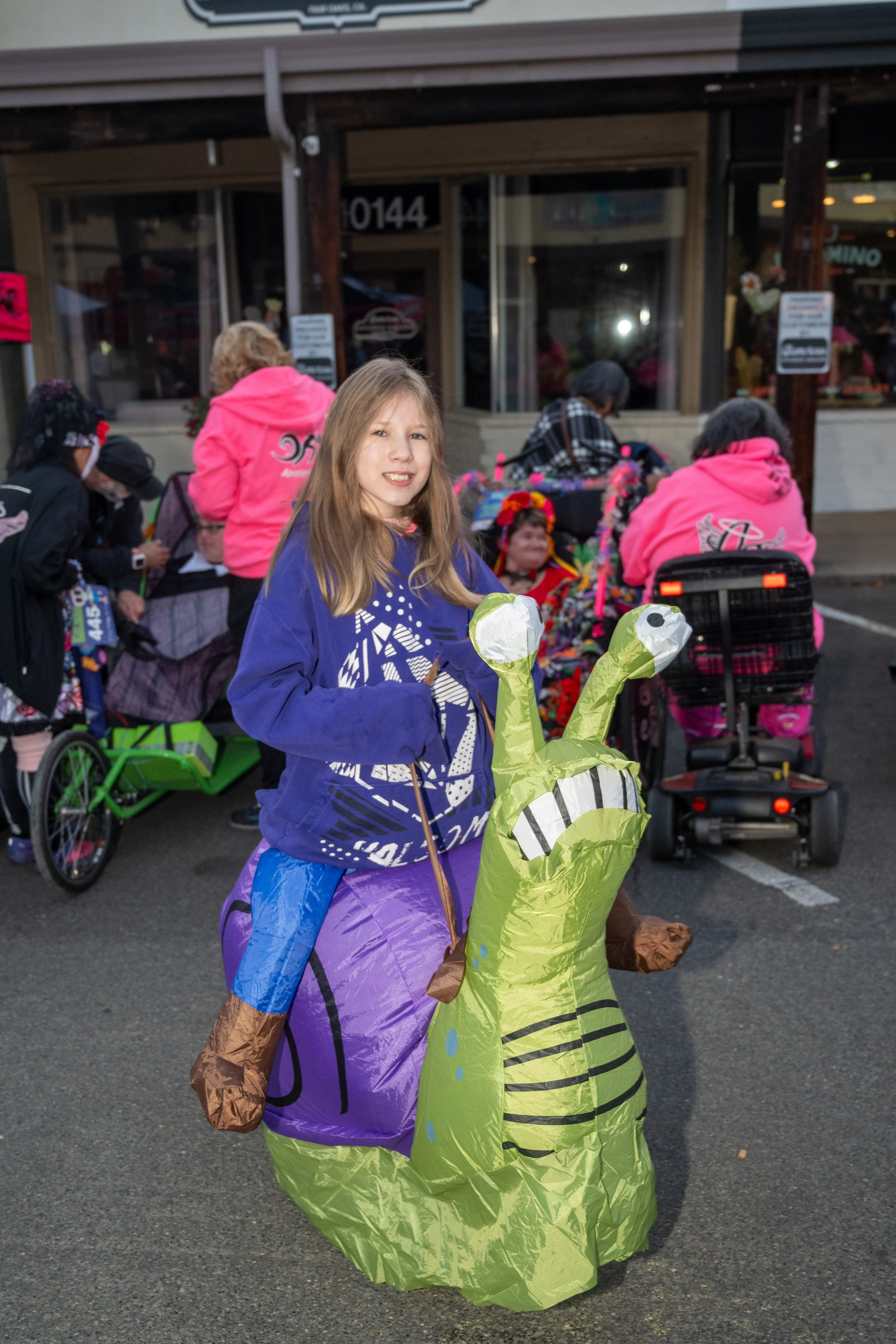 Young girl in a blue sweatshirt and purple pants riding a green inflatable dinosaur in a parking lot with adults and children dressed in costumes behind her.