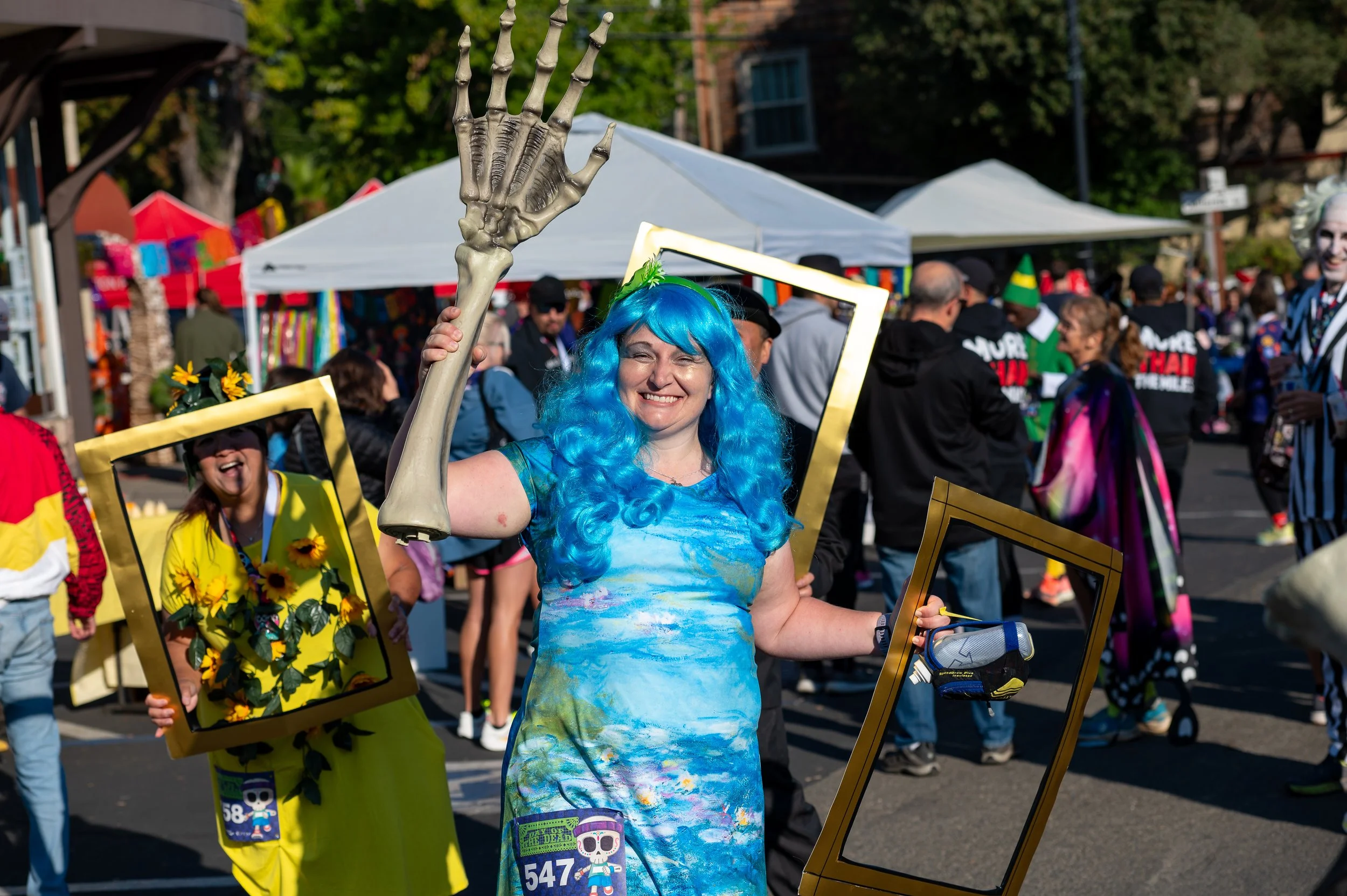 Woman with blue wig and tie-dye dress smiling at a colorful outdoor event, holding a skeleton arm prop and empty picture frames, with crowd and tents in the background.
