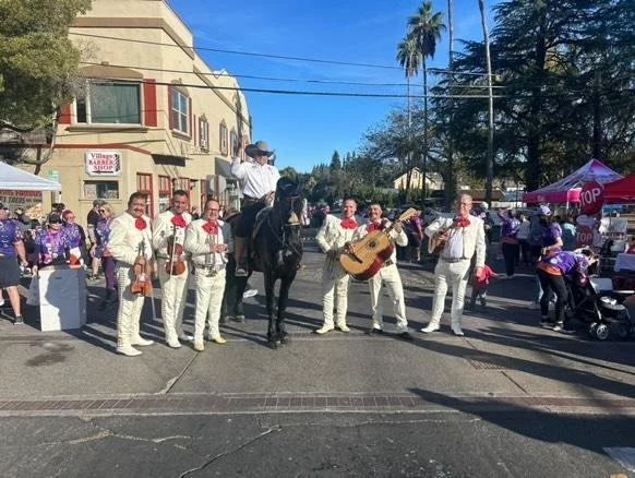 A mariachi band dressed in white suits with red bow ties performing on a street during a daytime festival, with a person on horseback and festival booths in the background.
