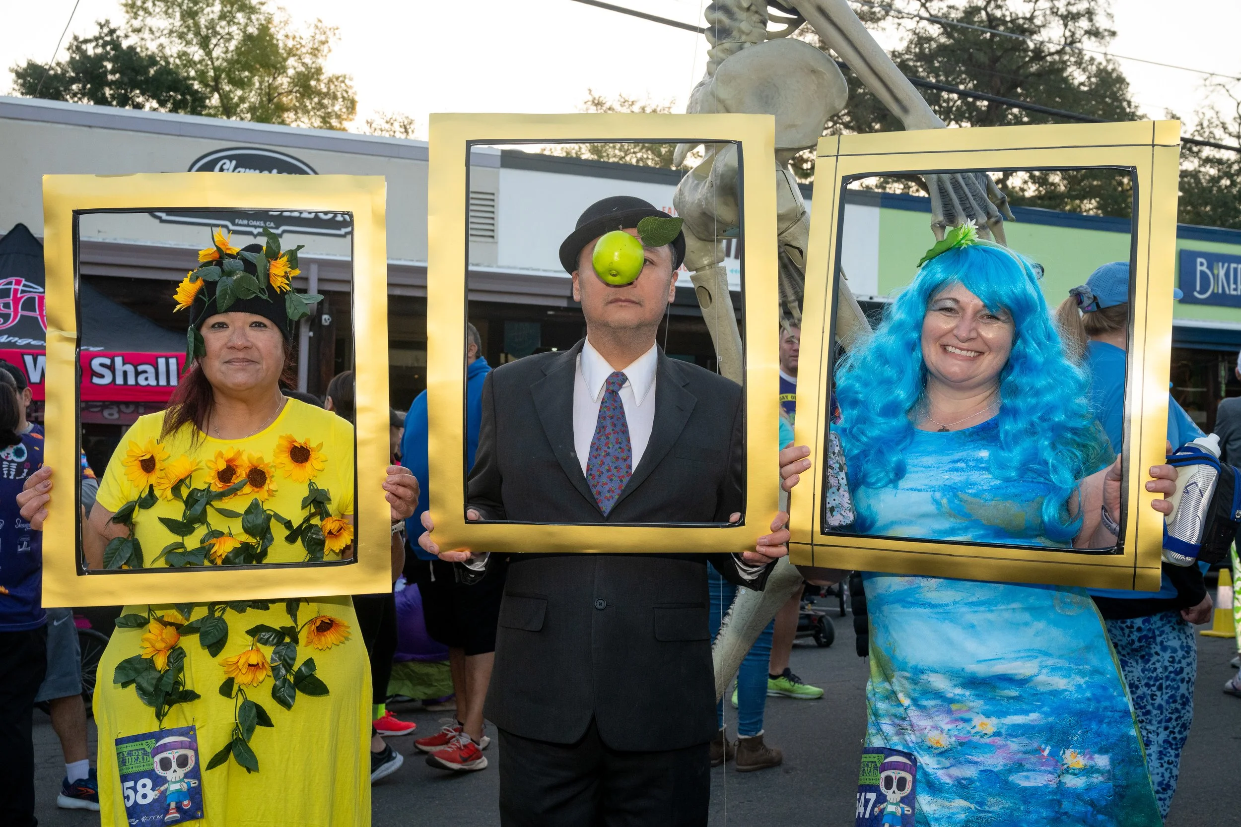 Three people dressed in colorful costumes holding picture frames, standing in an outdoor event. The person on the left is wearing a yellow dress decorated with sunflowers and a sunflower headpiece. The person in the middle is in a suit with a black b