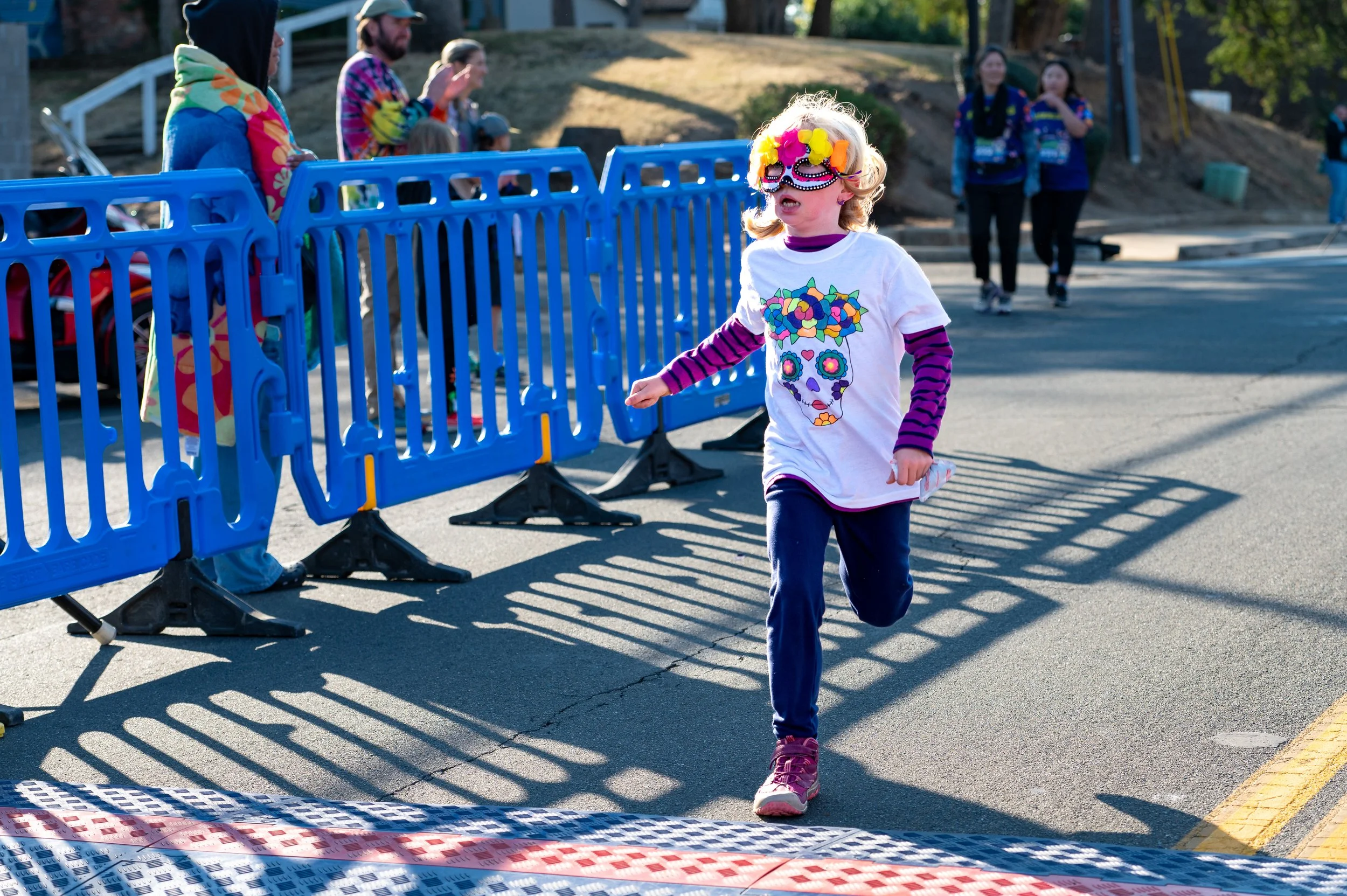 Young girl wearing a colorful sugar skull mask and a white T-shirt with a decorated skull design, running towards the finish line at a race.