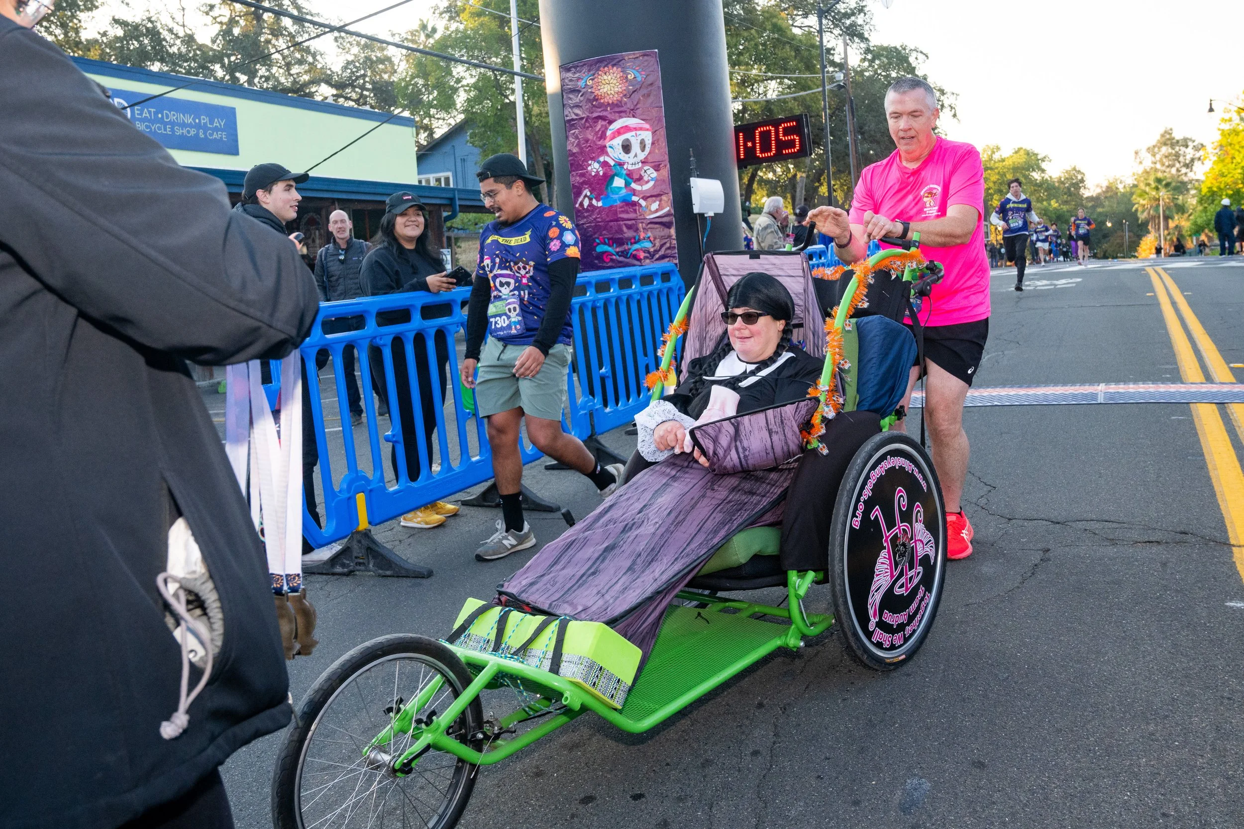 A woman in a wheelchair celebrating a race event, with a man pushing her, and other participants and spectators in the background on a city street.