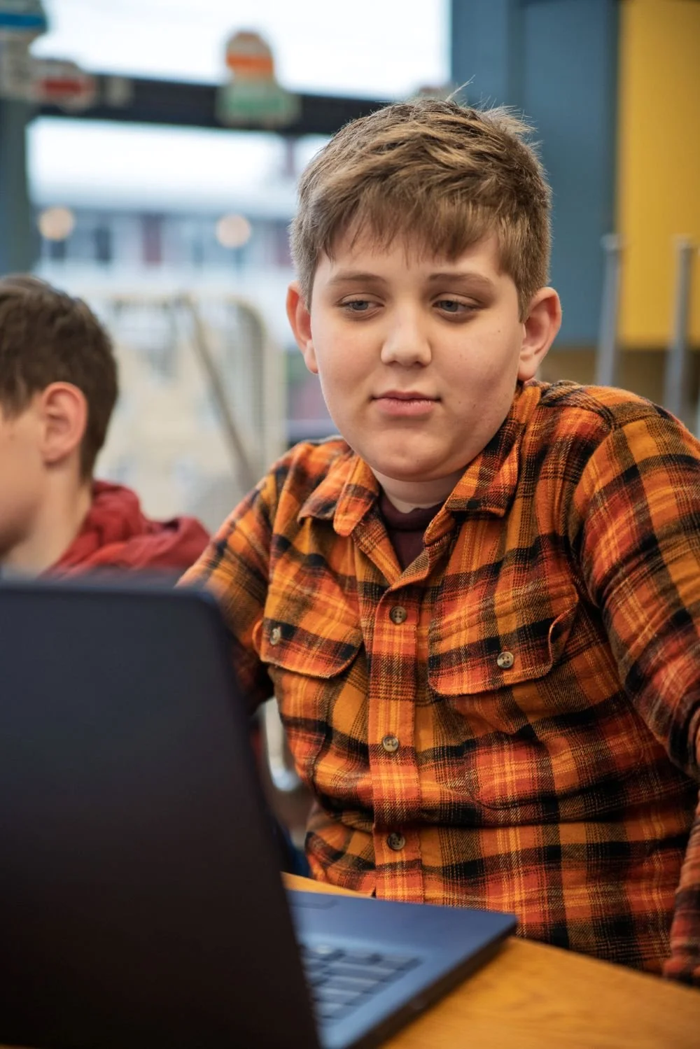 5th grade boy with plaid orange shirt and blonde hair looking at a laptop screen with a slight smile on his face