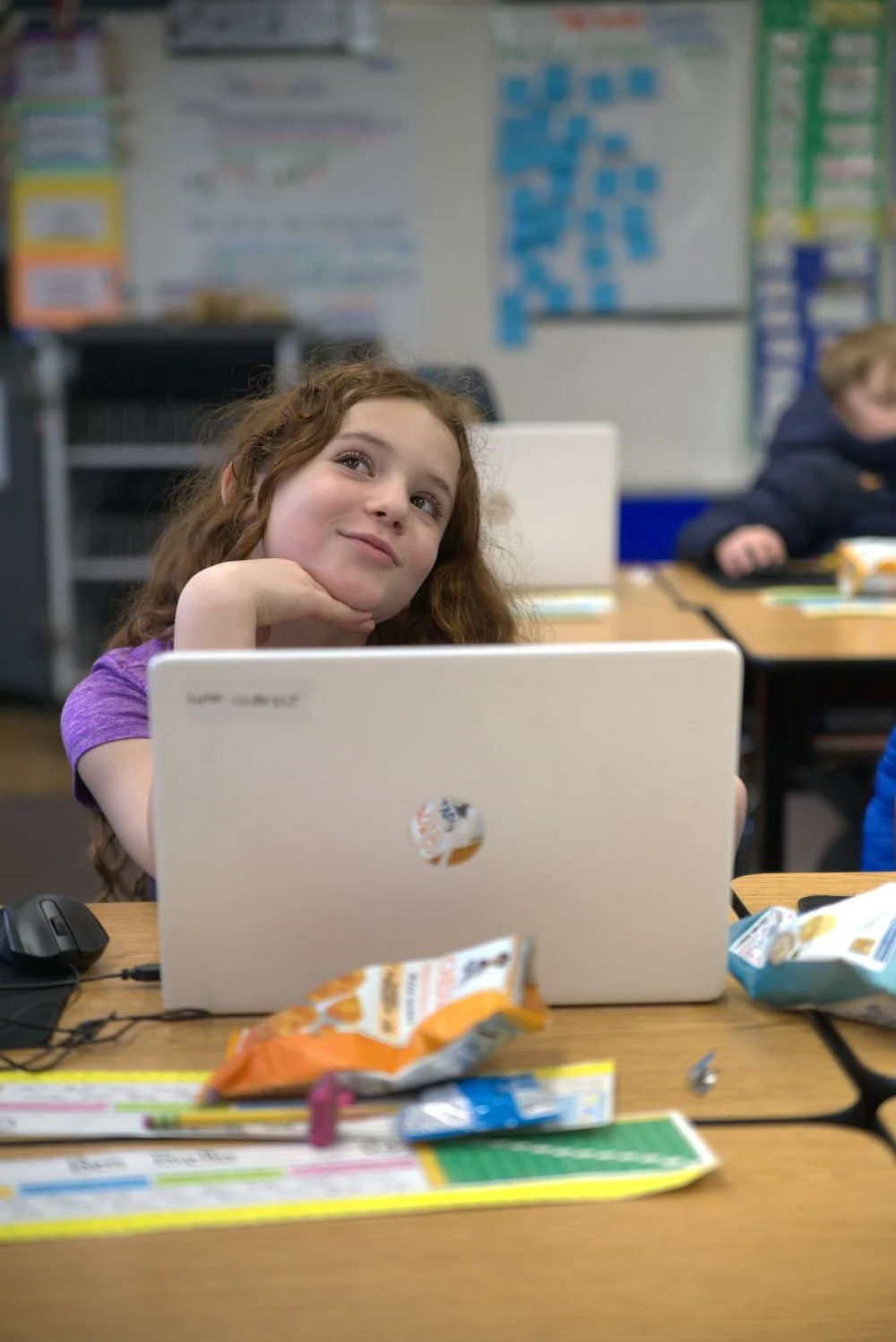 4th grade girl with light brown hair sitting behind an open laptop looking up and to the left in a cute pose with her fingers under her chin