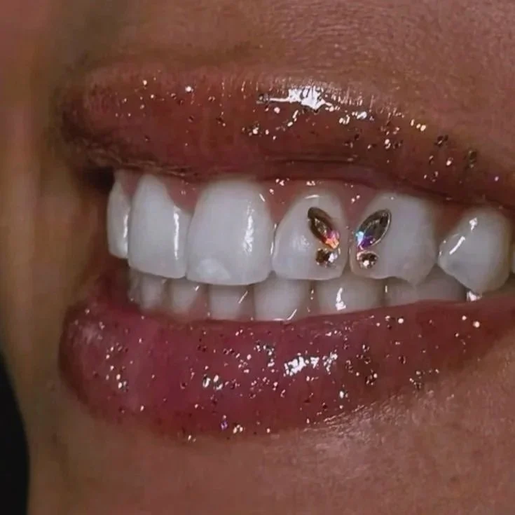 Close-up of a person's mouth showing white teeth with decorative rhinestone and colored studded lip piercings on the upper front tooth.