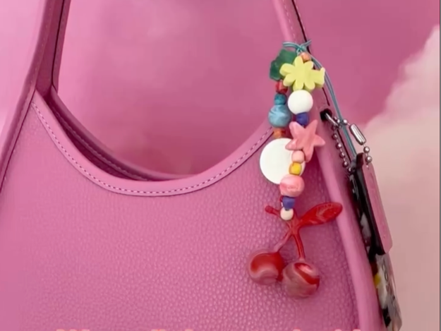 Close-up of a pink textured handbag with a colorful beaded strap and keychain hanging from it. The background is pink with some clouds.