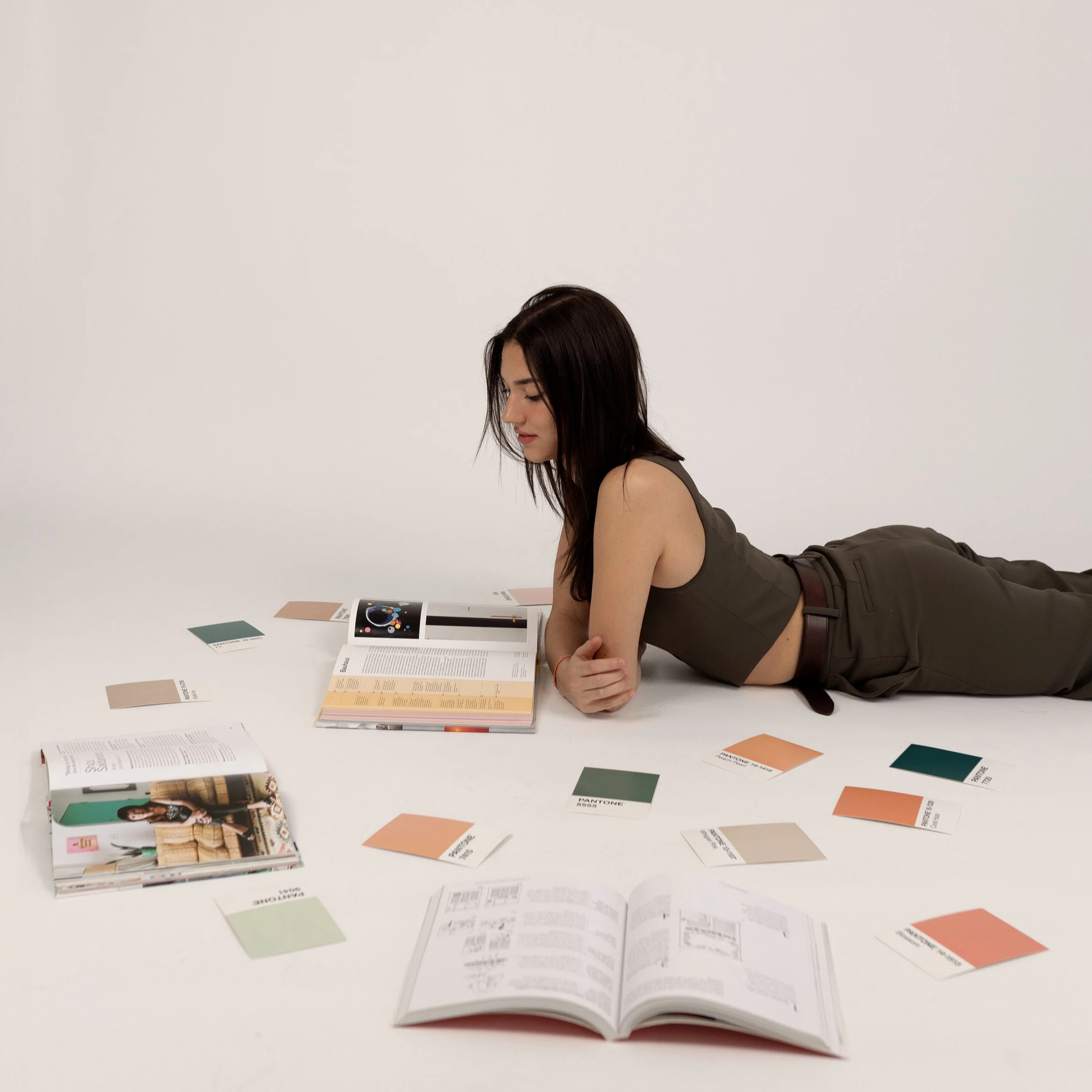 A woman lying on her stomach on a white surface, reading a magazine with color swatches and papers scattered around her. Milla V. Patricio