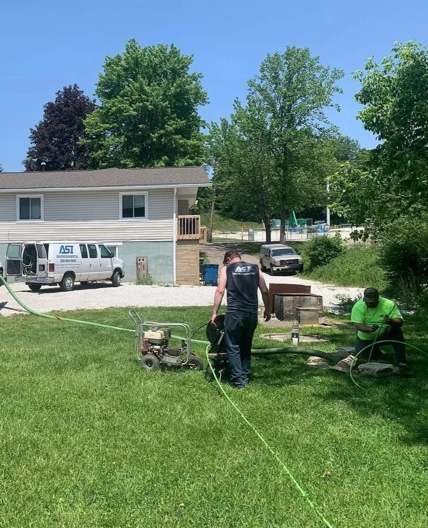 Two workers from ASI Environmental working outdoors on a lawn, one is operating equipment, the other is reading something on a mobile device, with houses, vehicles, and trees in the background on a sunny day.