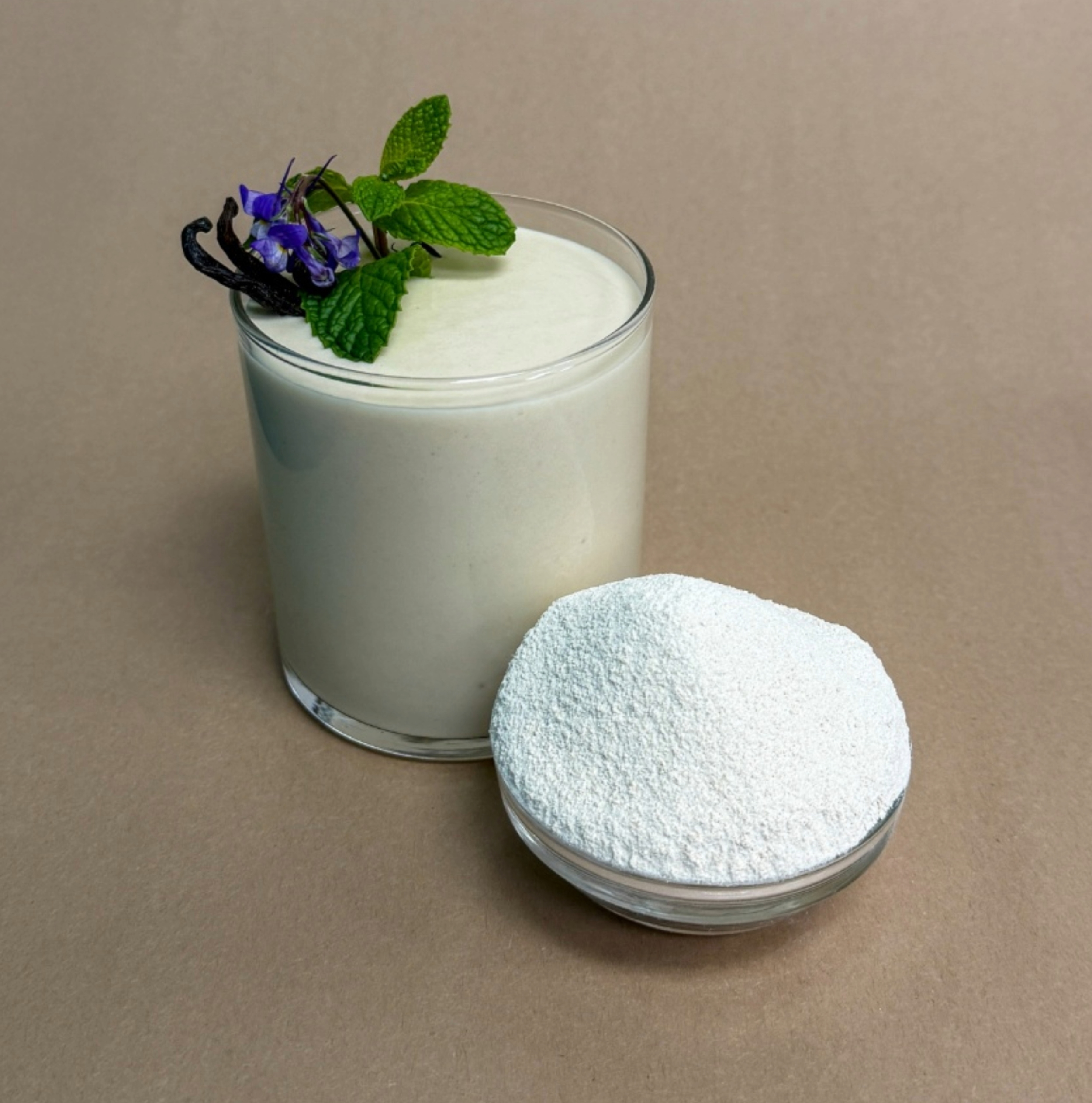 A glass of white smoothie garnished with mint leaves, purple flowers, and a vanilla bean, beside a small bowl of powdered protein powder on a beige background.