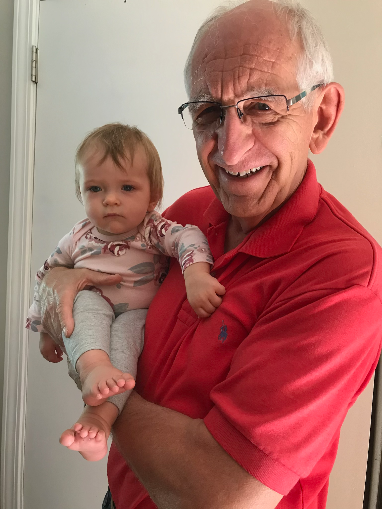 An elderly man with glasses holding a young girl in his arms. The man is smiling, wearing a red polo shirt, and the girl has a serious expression, wearing a floral top and gray pants.