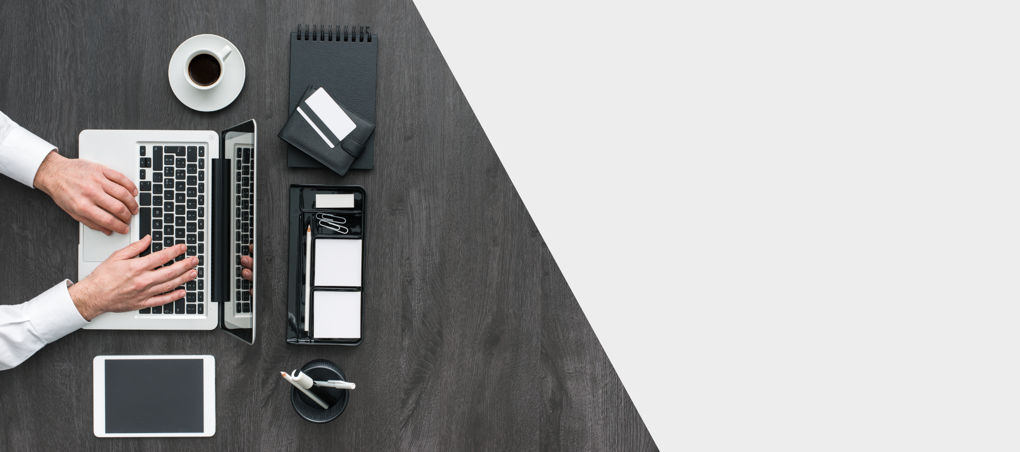 Overhead view of a workspace with a person's hands typing on a laptop, a cup of coffee in a saucer, a tablet, a black notebook with a small matching wallet, a black desk organizer with notepads, paper clips, a pen, and a pair of white earbuds on a dark wooden desk.
