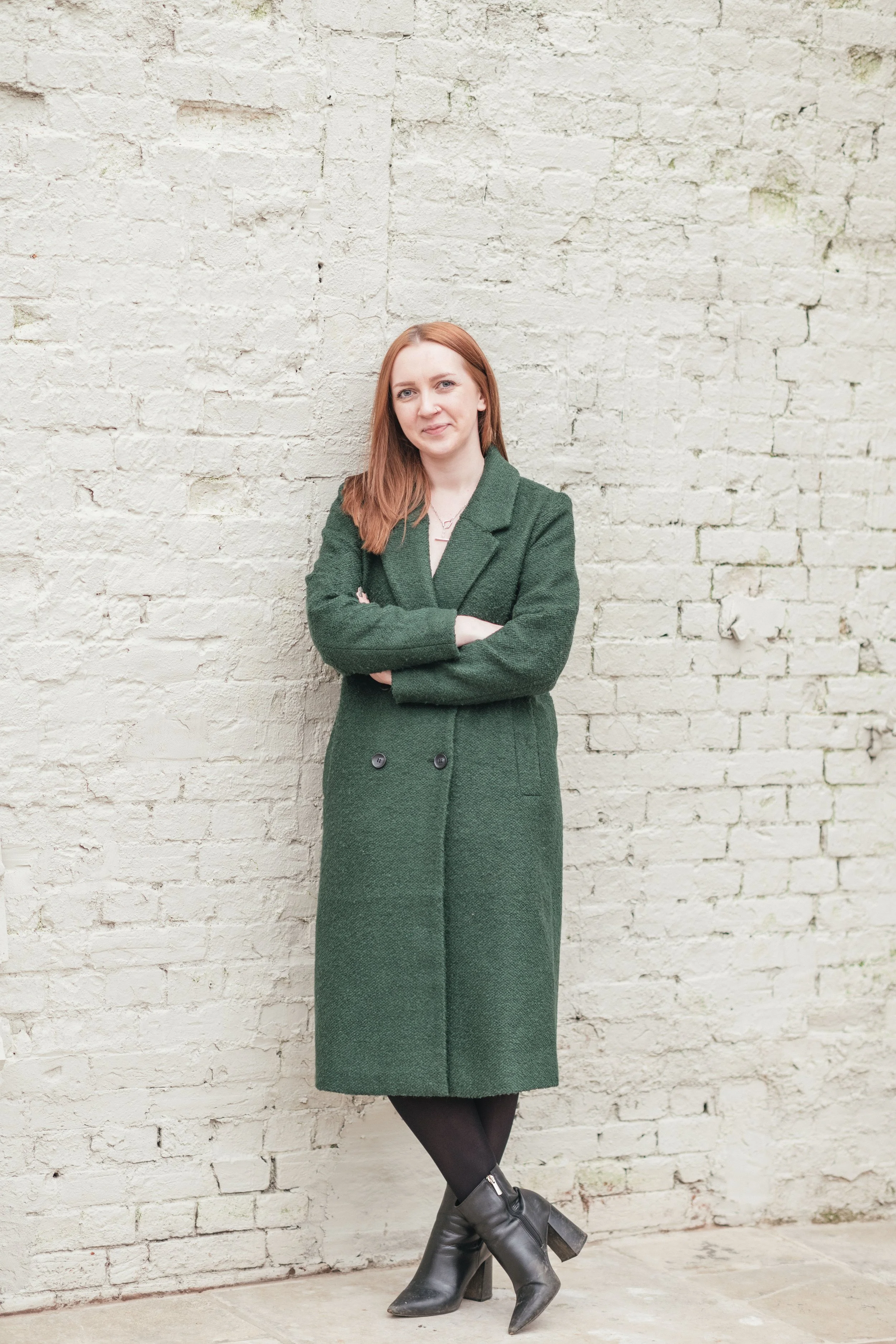 A woman with red hair wearing a green coat, black stockings, and black high-heeled boots standing with arms crossed in front of a white brick wall.