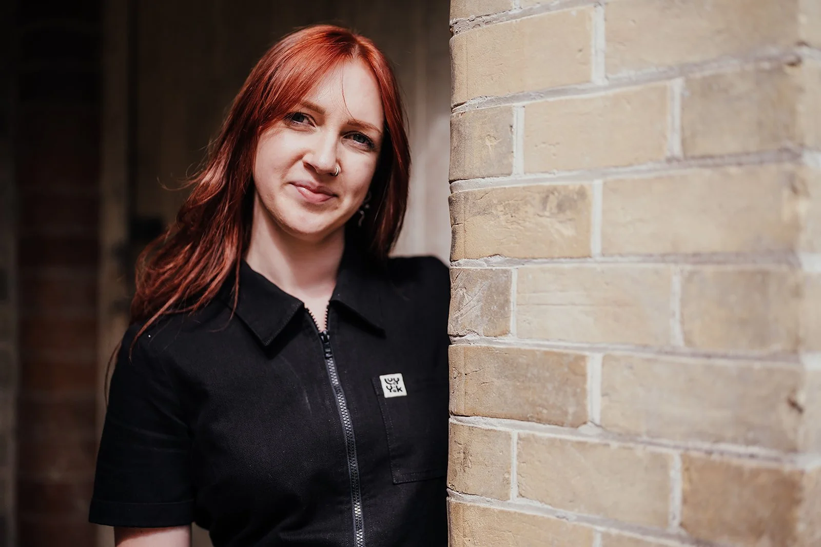 A woman with red hair and a nose piercing standing against a brick wall, wearing a black zip-up shirt and smiling.