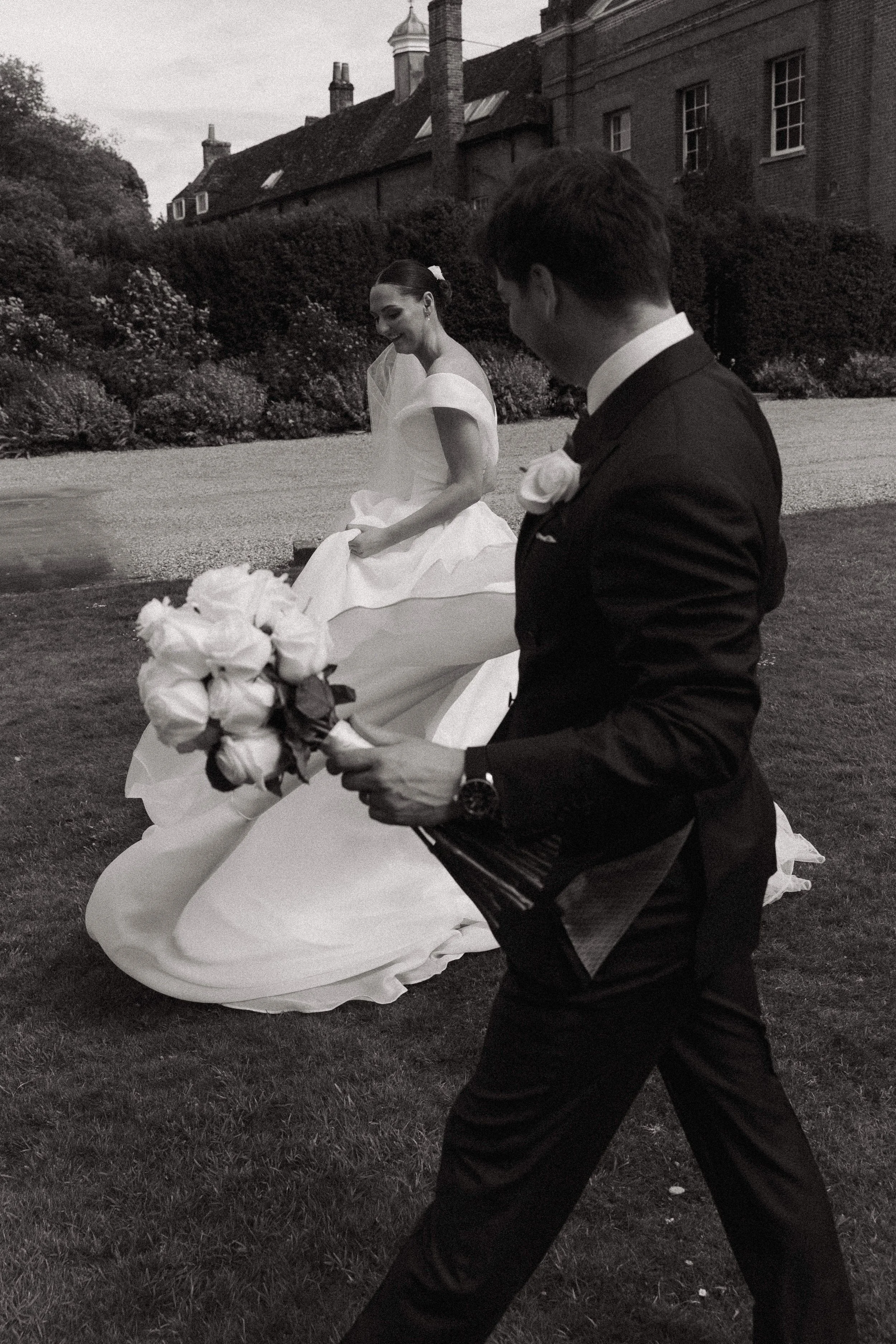 A black-and-white photograph of a groom walking with a bouquet, and a bride sitting on the grass in a garden with a background of an old brick building and blooming shrubs.