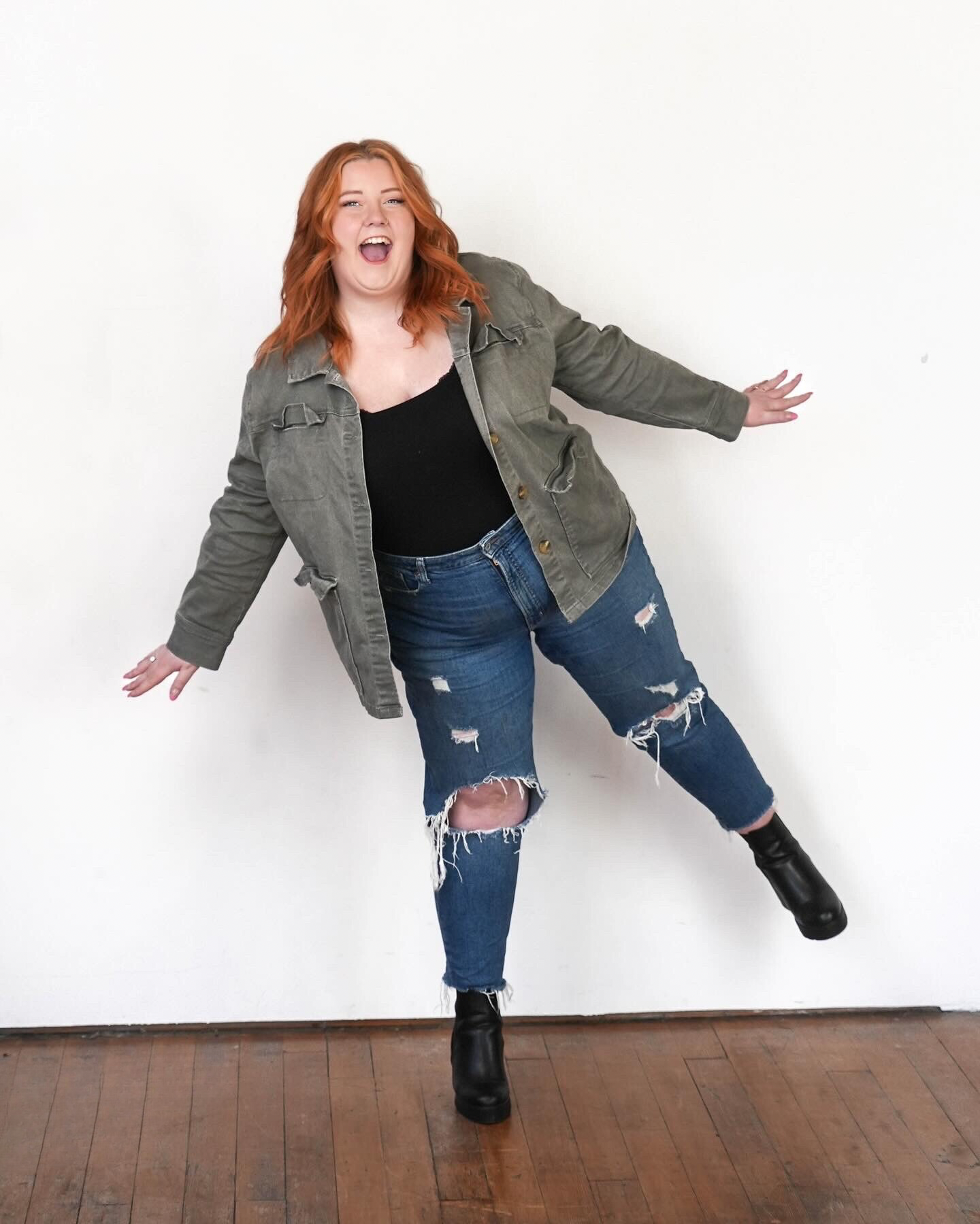 A woman with red hair, wearing a green jacket, black top, ripped jeans, and black boots, is smiling and balancing on one leg against a white wall.