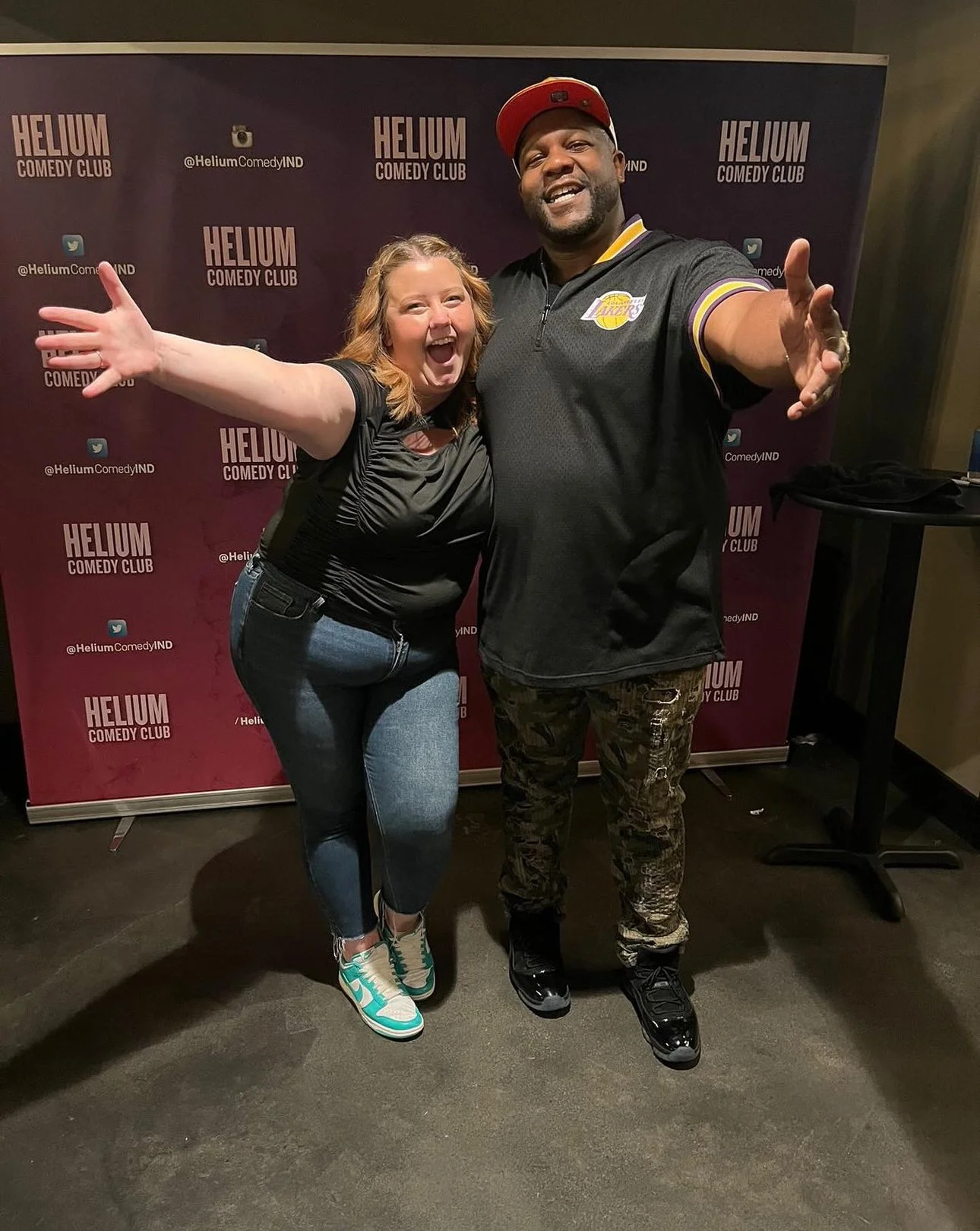 A woman and a man posing happily with their arms open in front of a Helium Comedy Club backdrop.