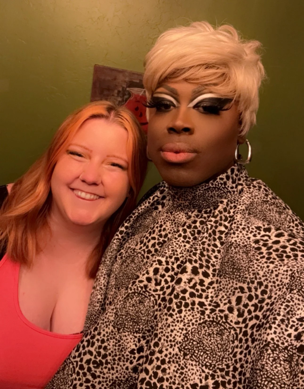 Two women posing for a selfie, one with red hair and the other with blonde hair and makeup, against a green wall.